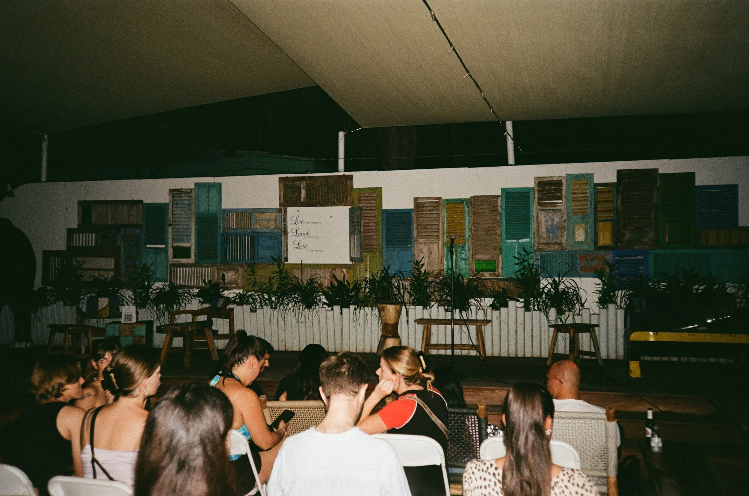 Audience sitting on chairs and benches in a dark indoor venue, watching a stage with a backdrop of mismatched wooden and painted shutters and potted plants.