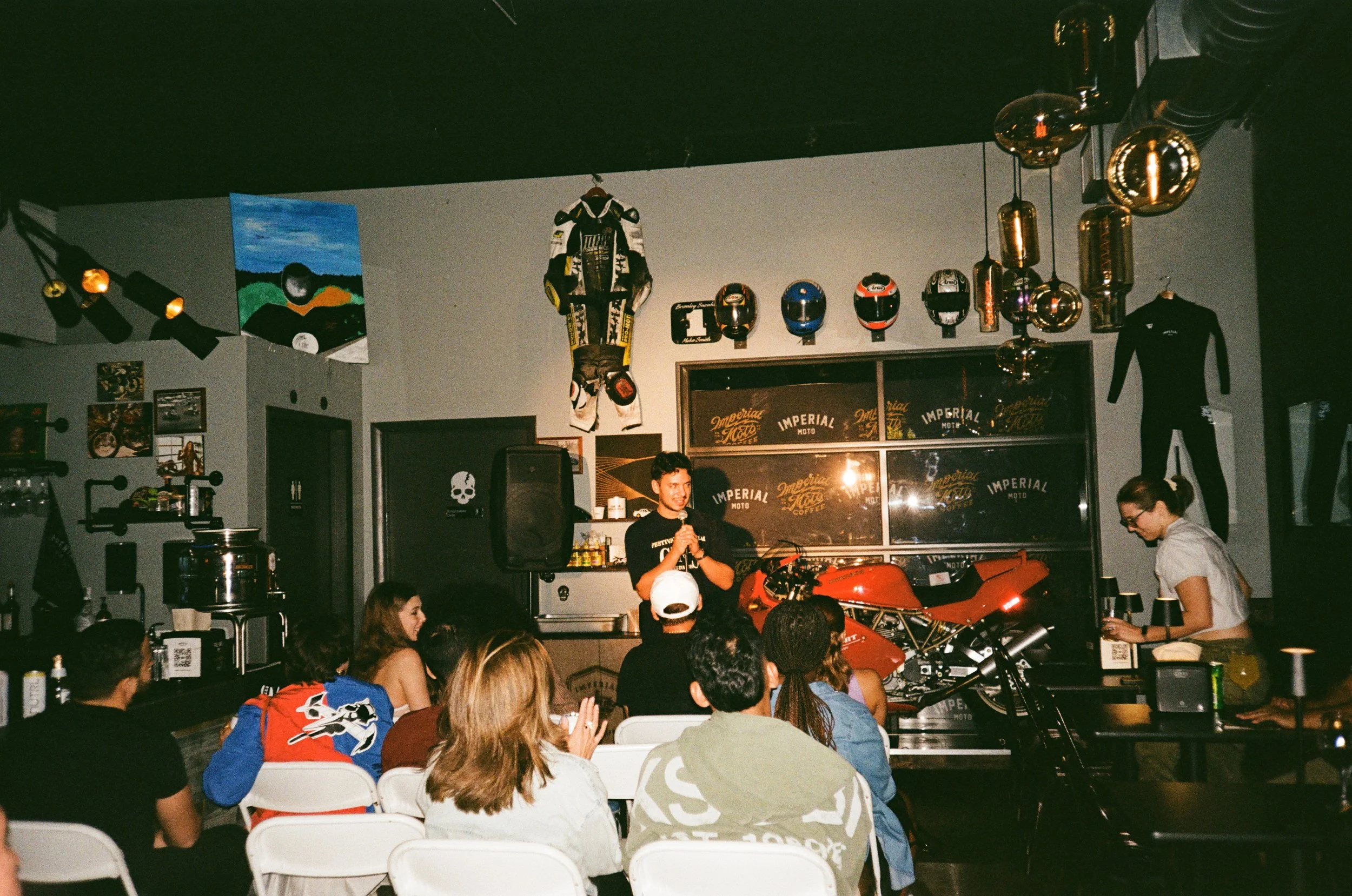 A group of people sitting at tables in a dimly lit cafe, listening to a young man speaking into a microphone on stage. The background features motorcycle-related decor, including helmets on the wall and a red motorcycle.