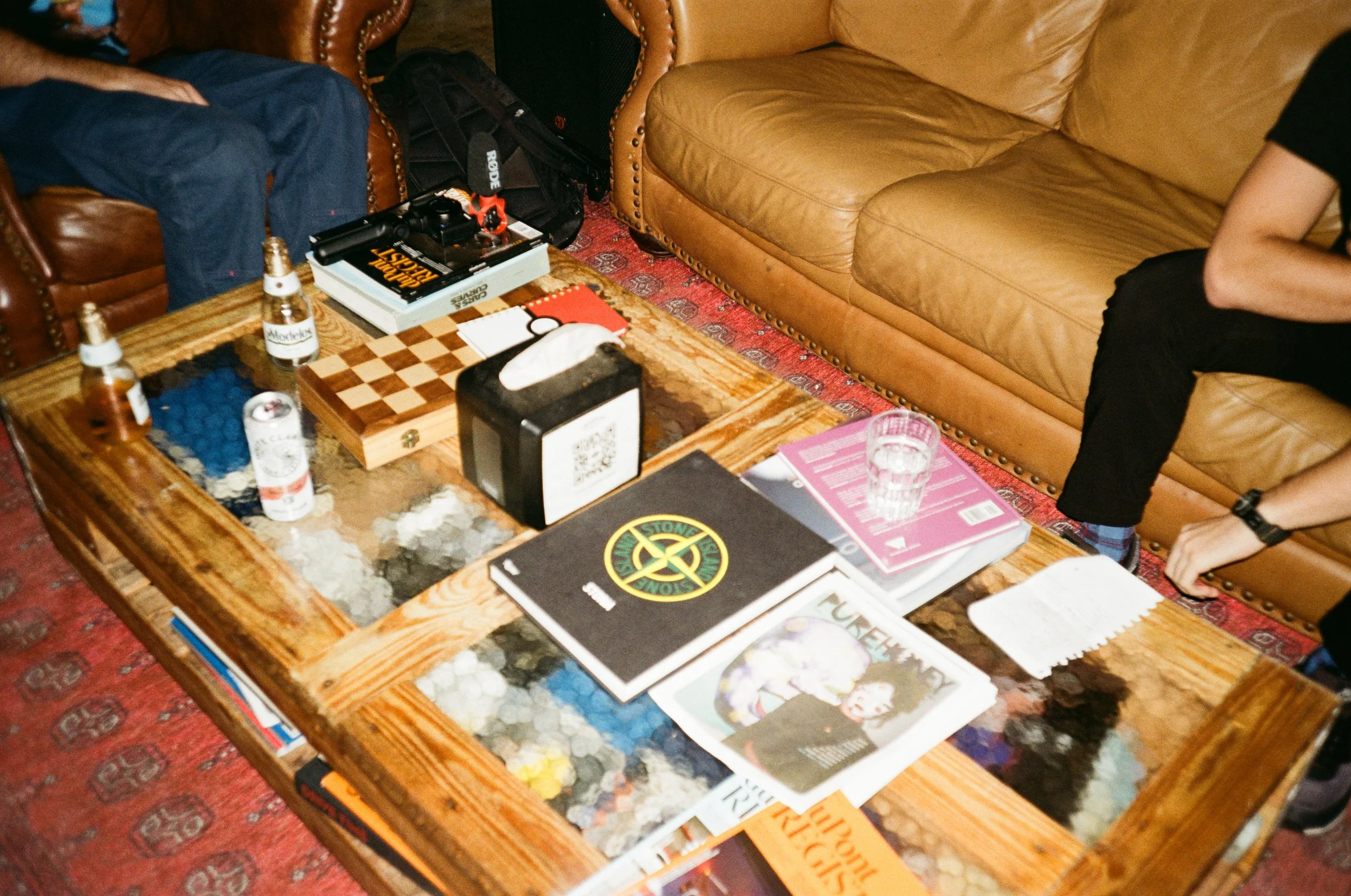 A wooden coffee table with a glass cover over colorful stones, surrounded by leather sofas and people partially visible sitting on them, with books, magazines, beer bottles, a tissue box, a glass of water, a chessboard, and a tissue box on the table.