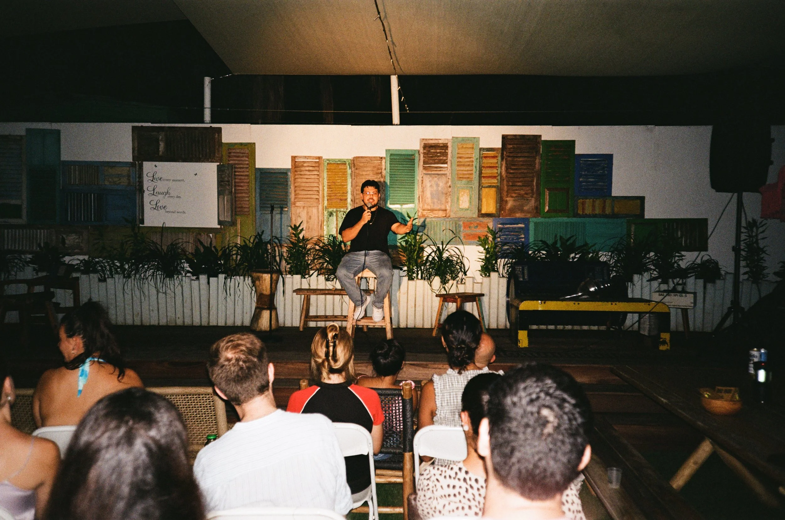 A man is sitting on a stool on a stage with a microphone, performing in front of an audience. Behind him, there are decorative wooden shutters and potted plants, with a sign that has the words 'Live,' 'Laugh,' and 'Love.' The audience is seated facin