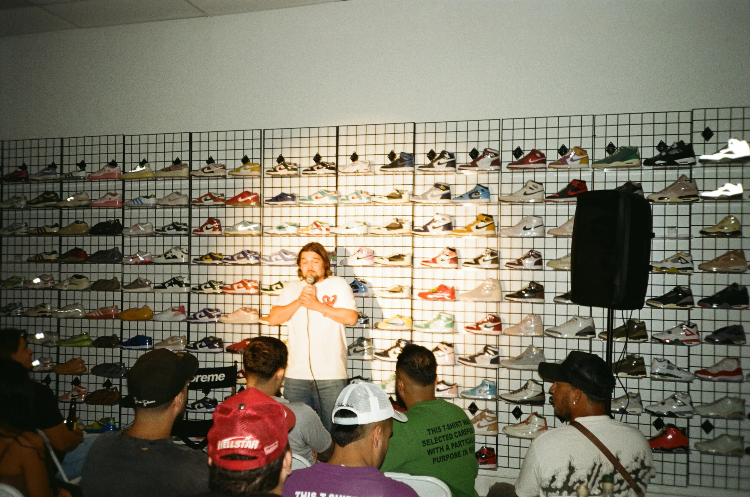 A person is speaking or singing into a microphone on stage with a large display of sneakers behind them. Several people are sitting and listening, with some wearing hats.