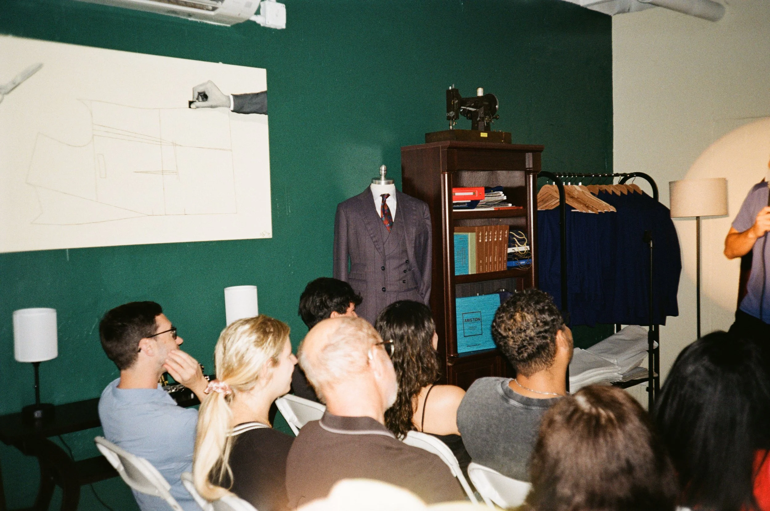 A group of people seated in chairs facing a presenter in a room decorated with a suit on a mannequin, a patch of yarn, and books on a shelf, with a whiteboard displaying a sketch of a shape.