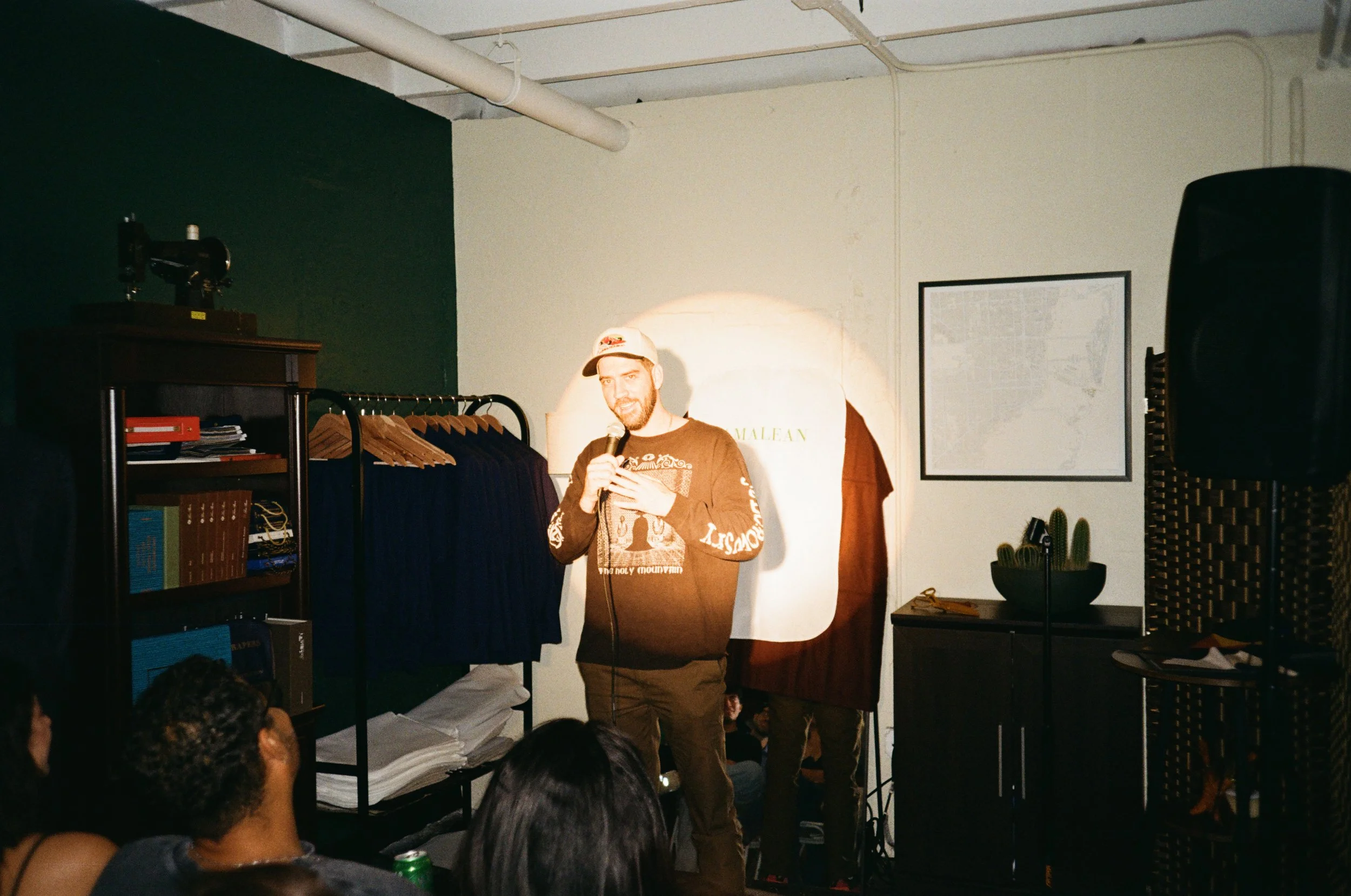 A man with a beard, wearing a beige baseball cap and a dark long-sleeve shirt, is speaking into a microphone during a stand-up comedy performance. He is standing in front of a white wall illuminated by a spotlight, with an audience seated in front of