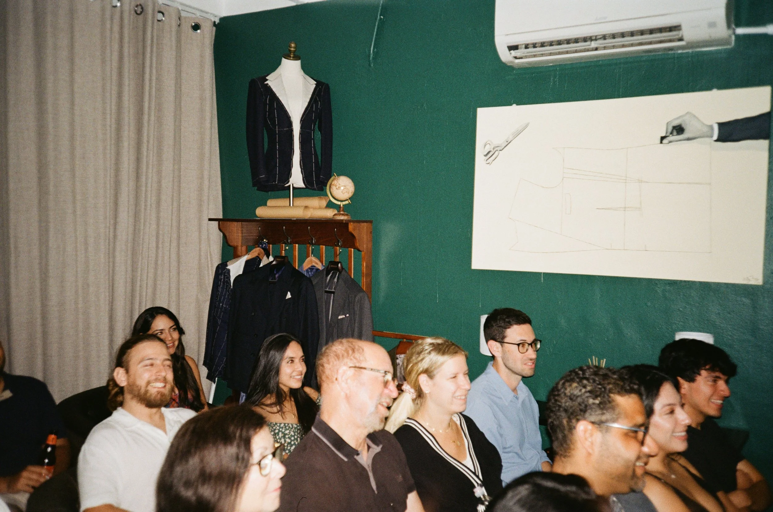 A group of people sitting and smiling in a room with green walls and decor, including a coat rack with jackets, a small globe, and a large piece of wall art with a scarecrow, scissors, and sewing lines.