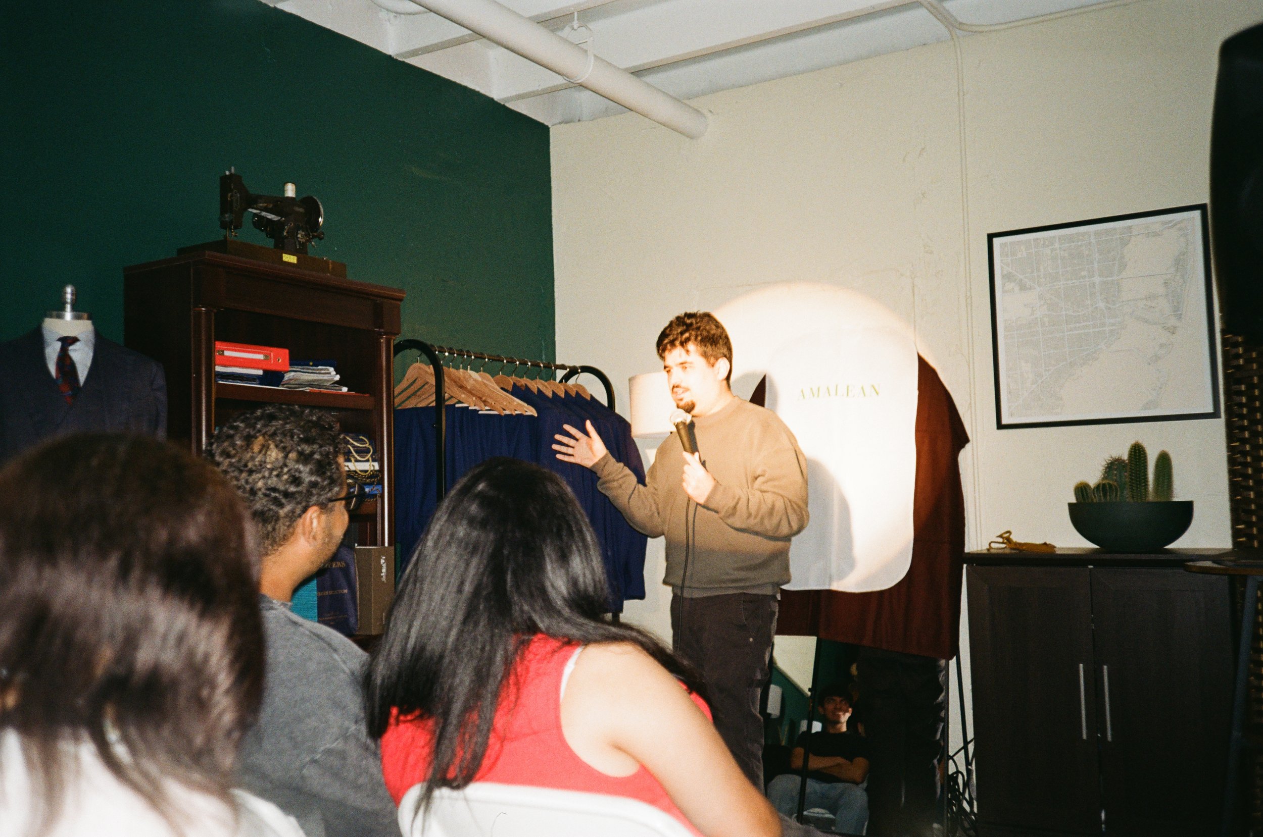 A man giving a presentation or speech to an audience in a room decorated with clothing racks, a map, and plants, with a small group of people seated and listening.
