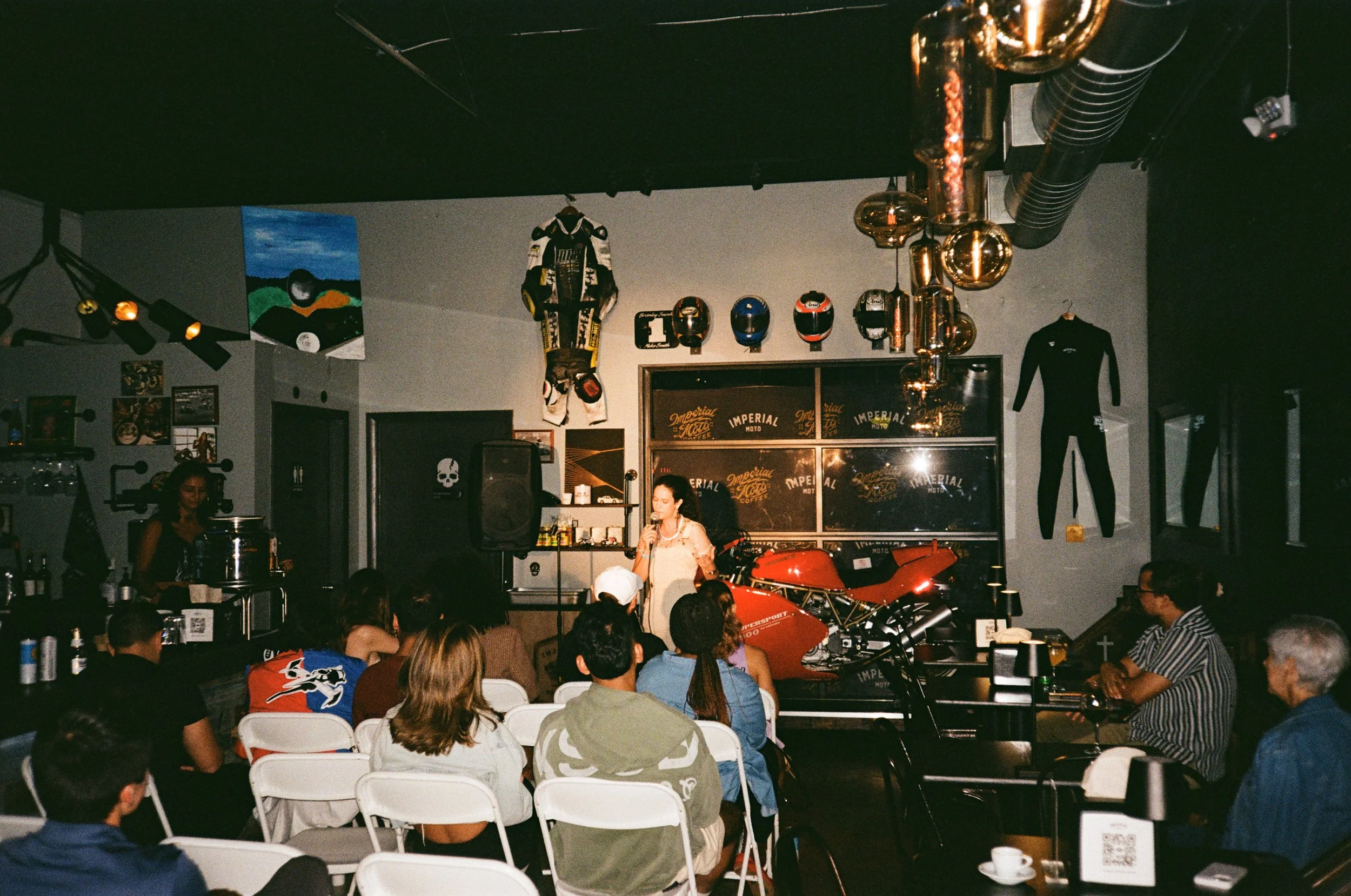 An indoor event space with a woman speaking into a microphone in front of a red motorcycle. The decor includes racing helmets, racing suits, artwork, and automotive-themed items on the wall, with an audience seated and watching.