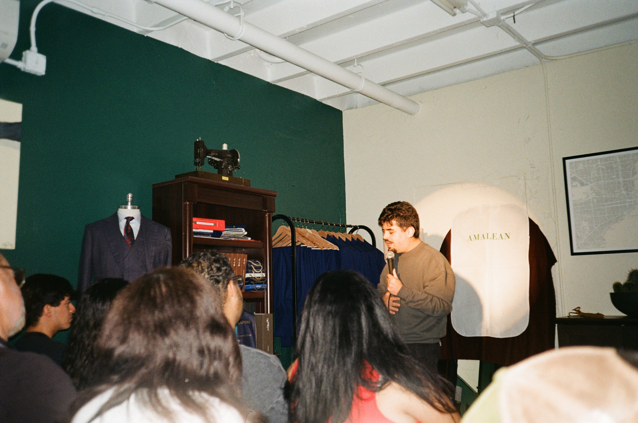 A young man giving a presentation, holding a microphone, in front of an audience. The setting appears to be a classroom or seminar room with clothing and vintage items on display, including a suit on a mannequin and a cabinet with books and a sewing 