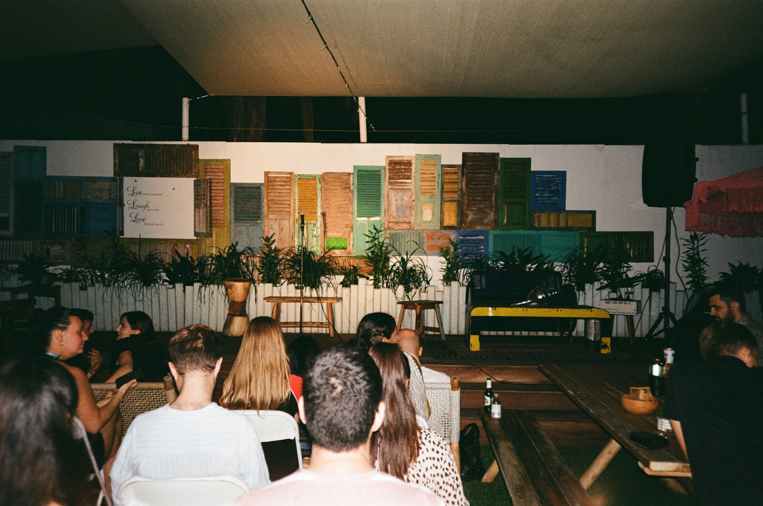 People seated in a dimly lit room with a stage decorated with colorful, vintage shutters and plants in front, prepared for a performance or event.