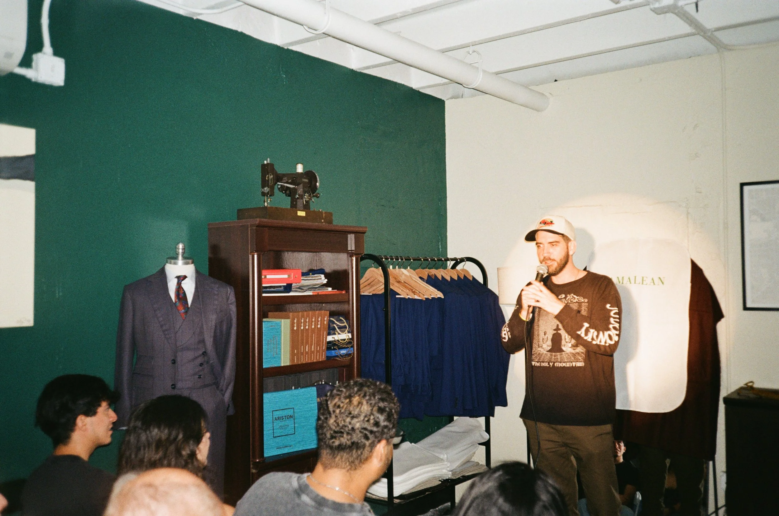 A man standing with a microphone in hand speaking to an audience in a room decorated with vintage items, including a mannequin dressed in a suit, a bookshelf with books, a clothing rack with blue garments, and a large framed photo on the wall.