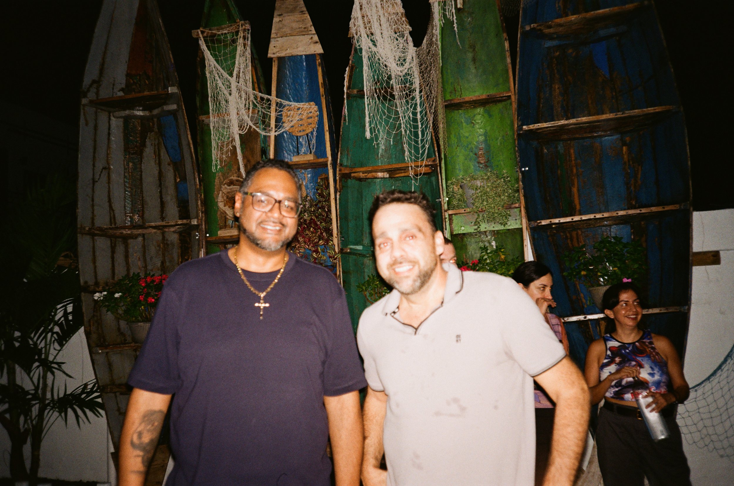 Two men smiling in front of a colorful wooden backdrop with plants and fishing nets, at a social gathering.