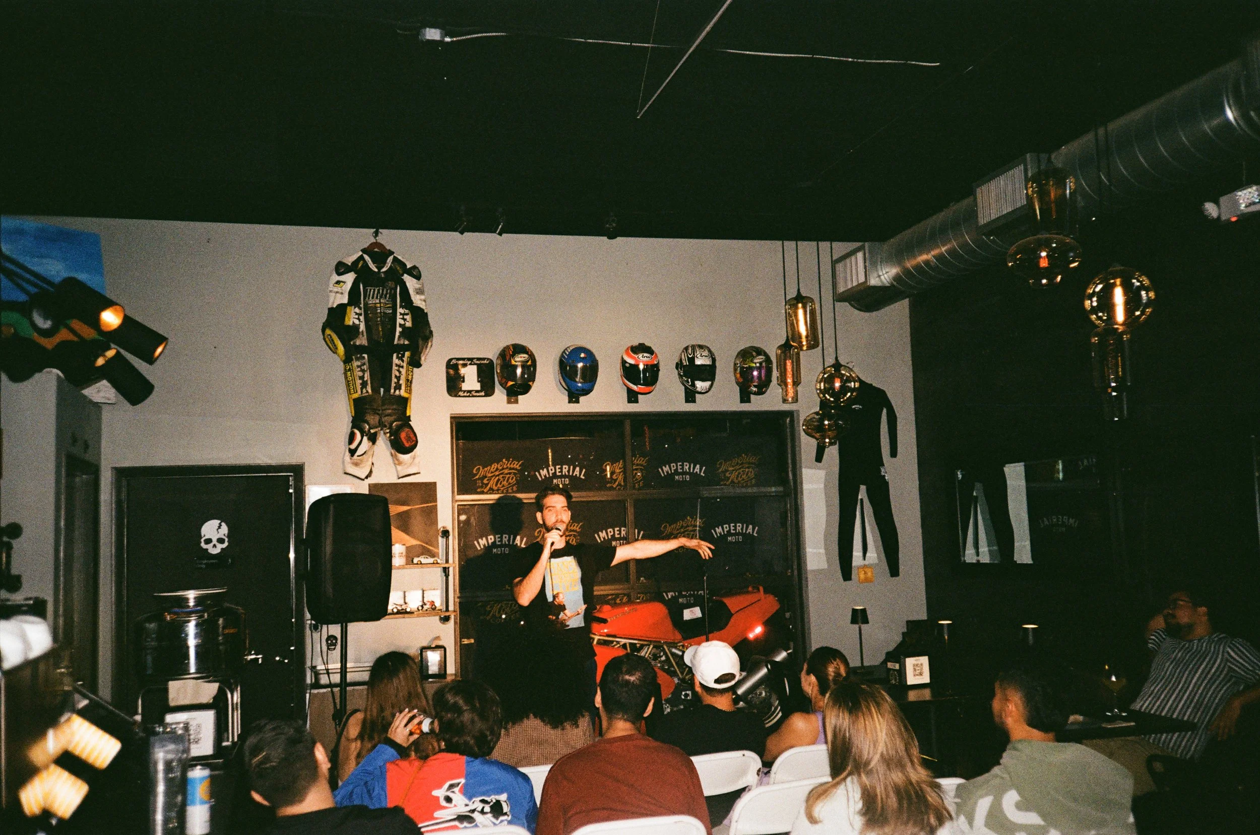 A group of people sitting and listening to a speaker on a stage in a room with motorcycle helmets and gear on the wall.