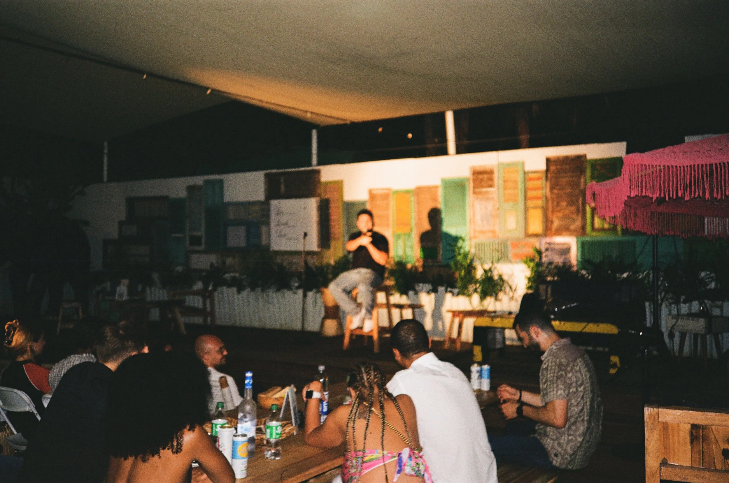 People sitting at a table watching a performer on a small stage in a dimly lit venue.