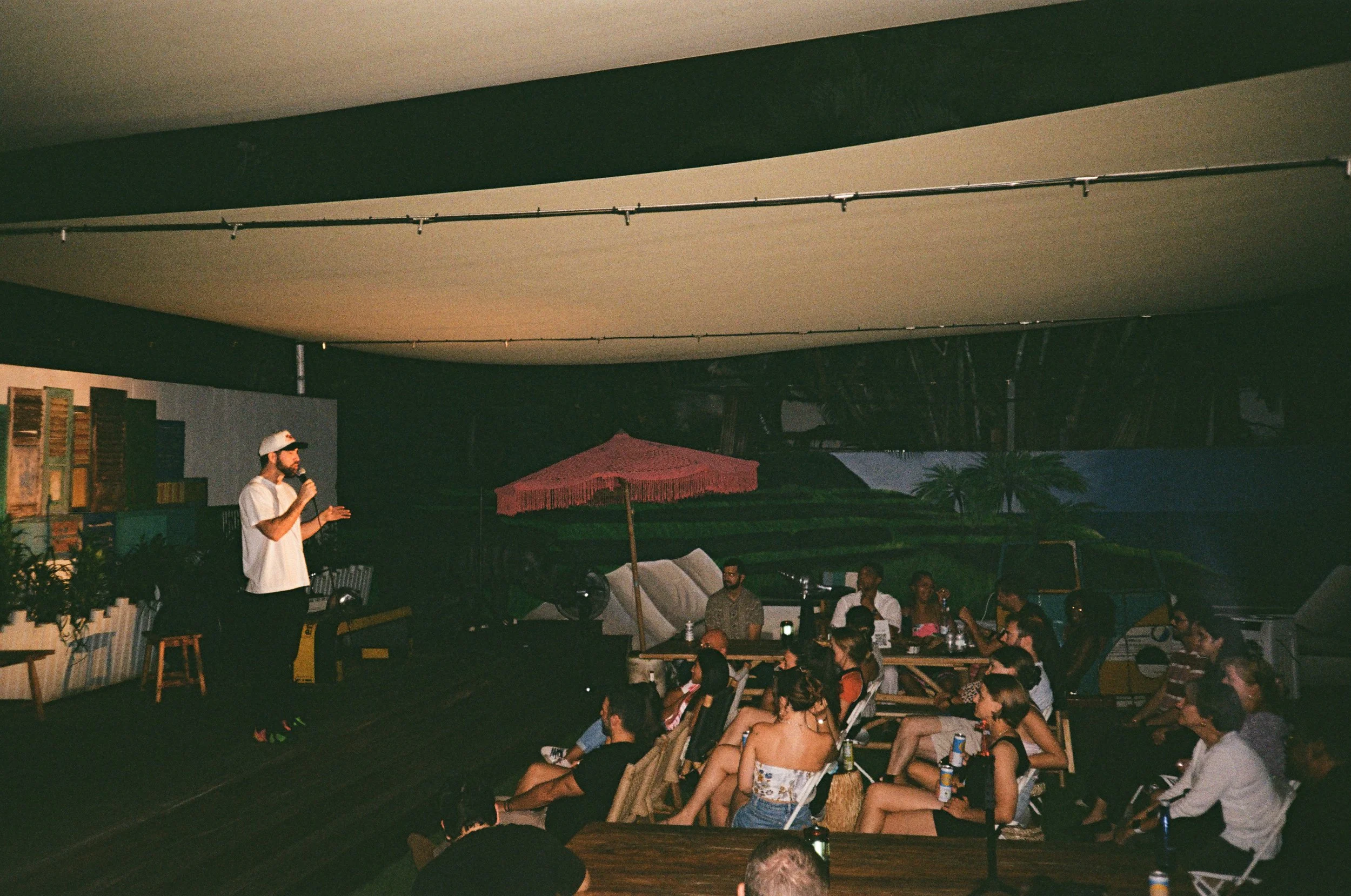 A man with a beard in a white shirt and baseball cap speaks into a microphone on a small stage, while an audience of young adults sits on outdoor furniture, listening during nighttime.