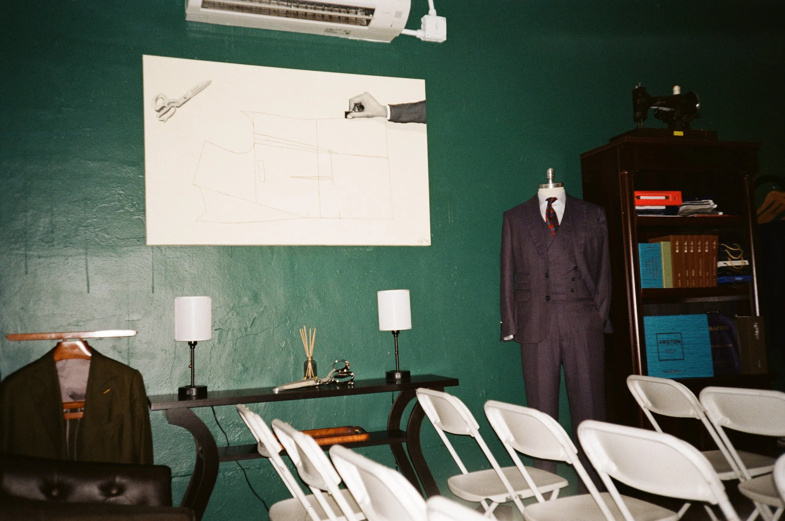 Interior of a room with white folding chairs, a dark wood cabinet, a mannequin dressed in a suit, a wall with a large sketch, and a table with lamps and accessories.