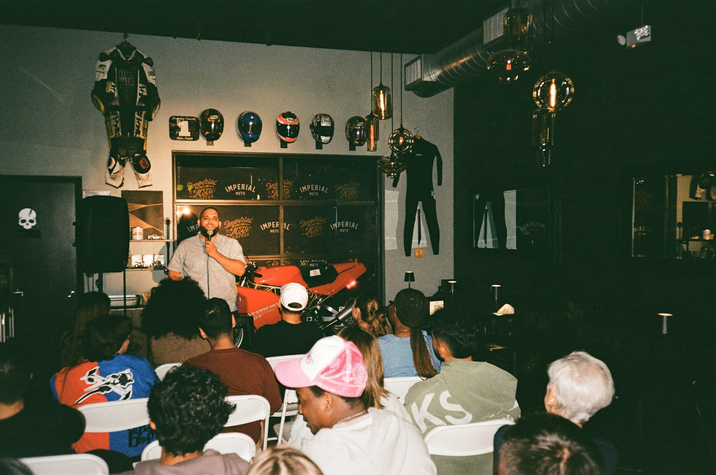 A man with a beard holding a microphone performs stand-up comedy on stage in a dimly lit venue. The audience sits in white chairs, and the decor includes racing helmets, motorcycle gear, and a red motorcycle.