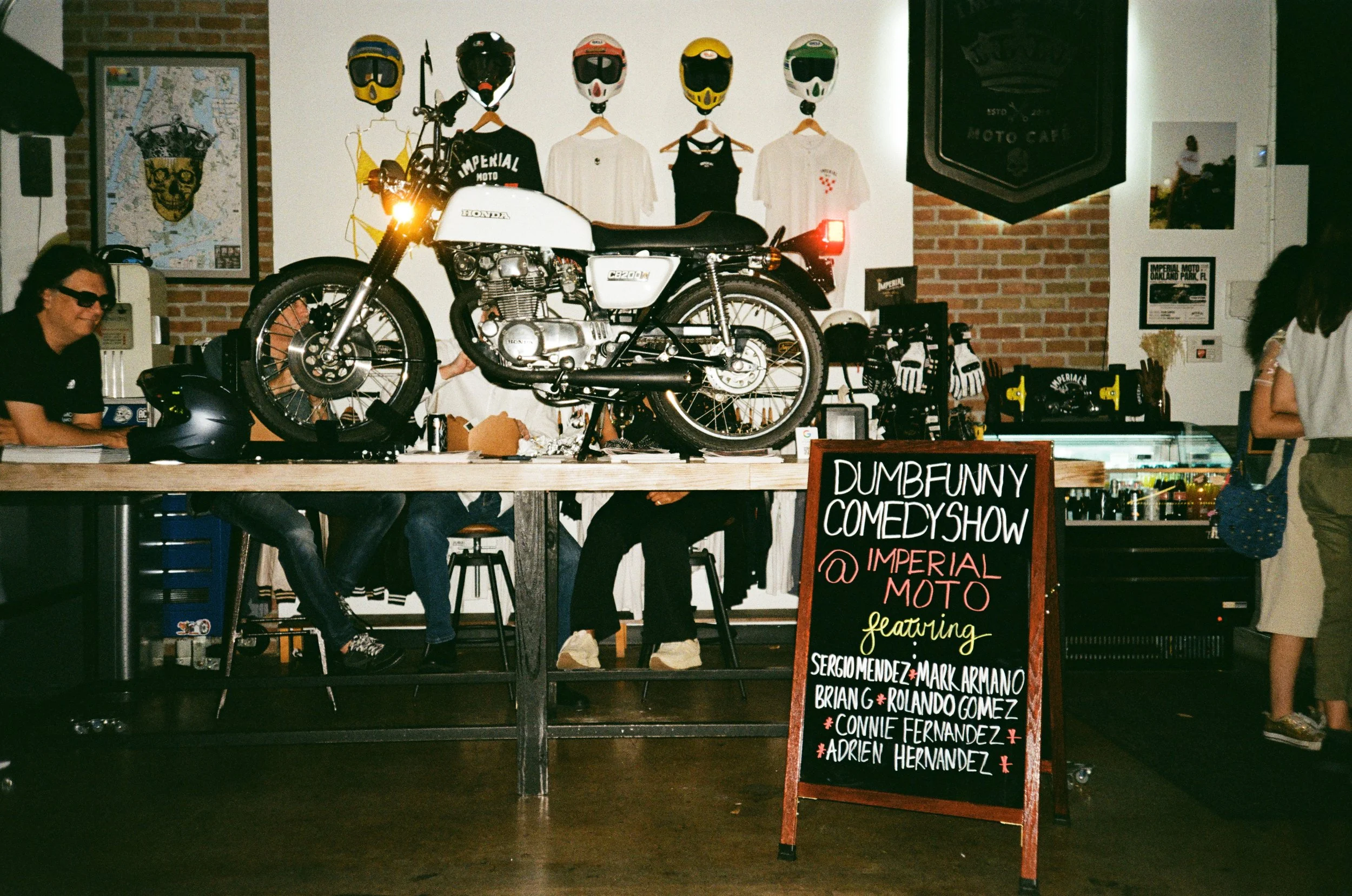 People sitting on stools at a bar with a vintage Honda motorcycle displayed on the countertop. The background features helmets, shirts, and posters related to motorcycles at the Imperial Moto cafe, advertising a comedy show featuring several performe