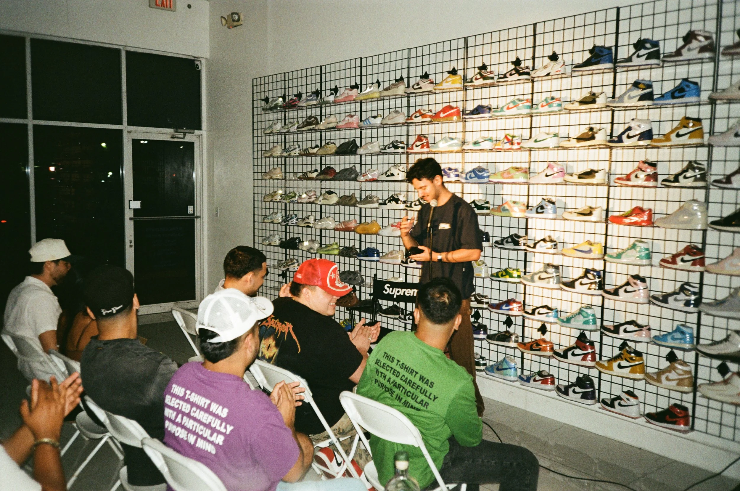 Group of people sitting on chairs in front of a wall display of sneakers, with a man speaking into a microphone.