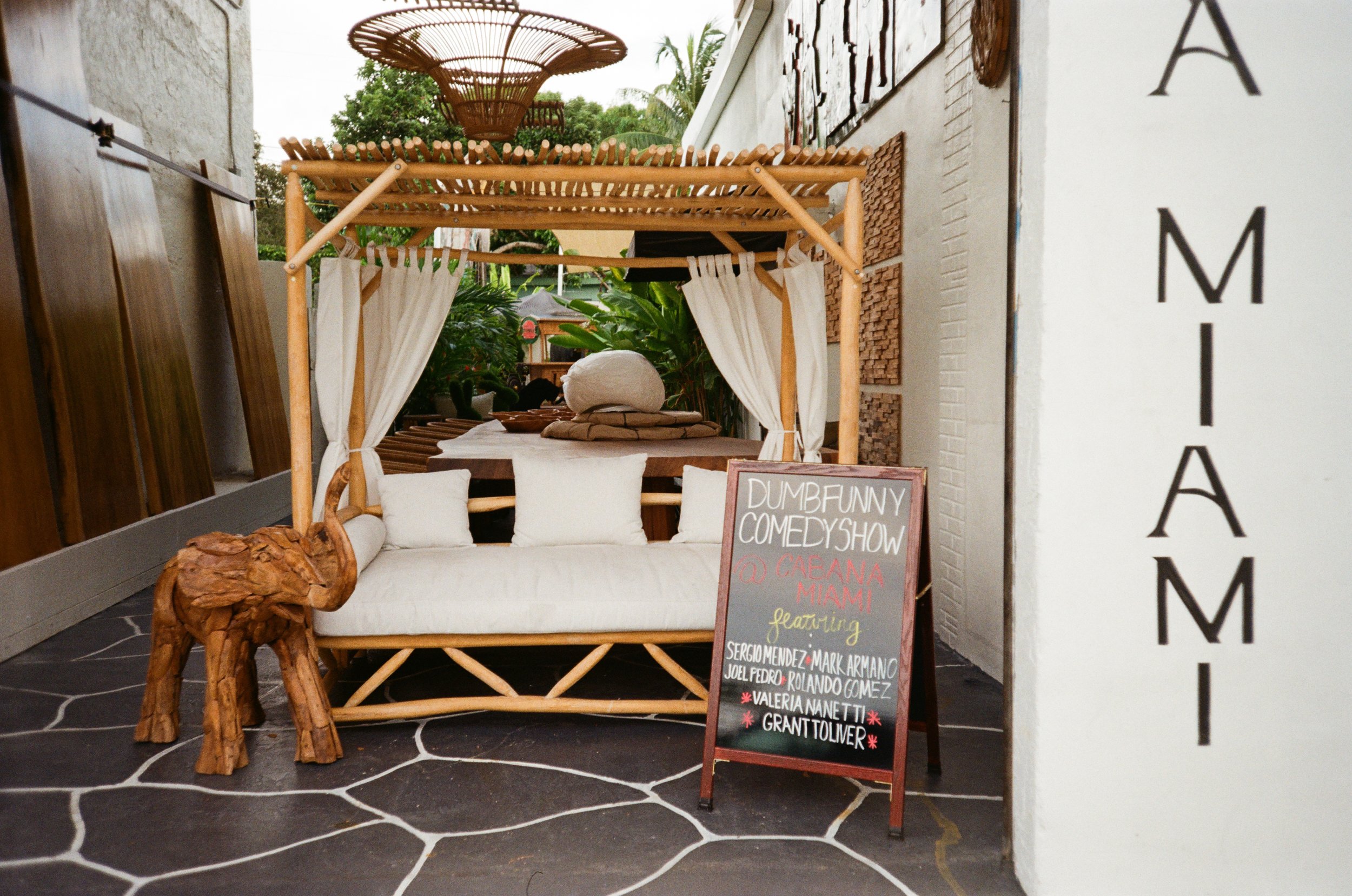 Outdoor area featuring a bamboo canopy with white curtains, a wooden sofa with cushions, a small wooden table, a carved wooden elephant, a chalkboard sign with comedy show details, and lush green plants in the background.