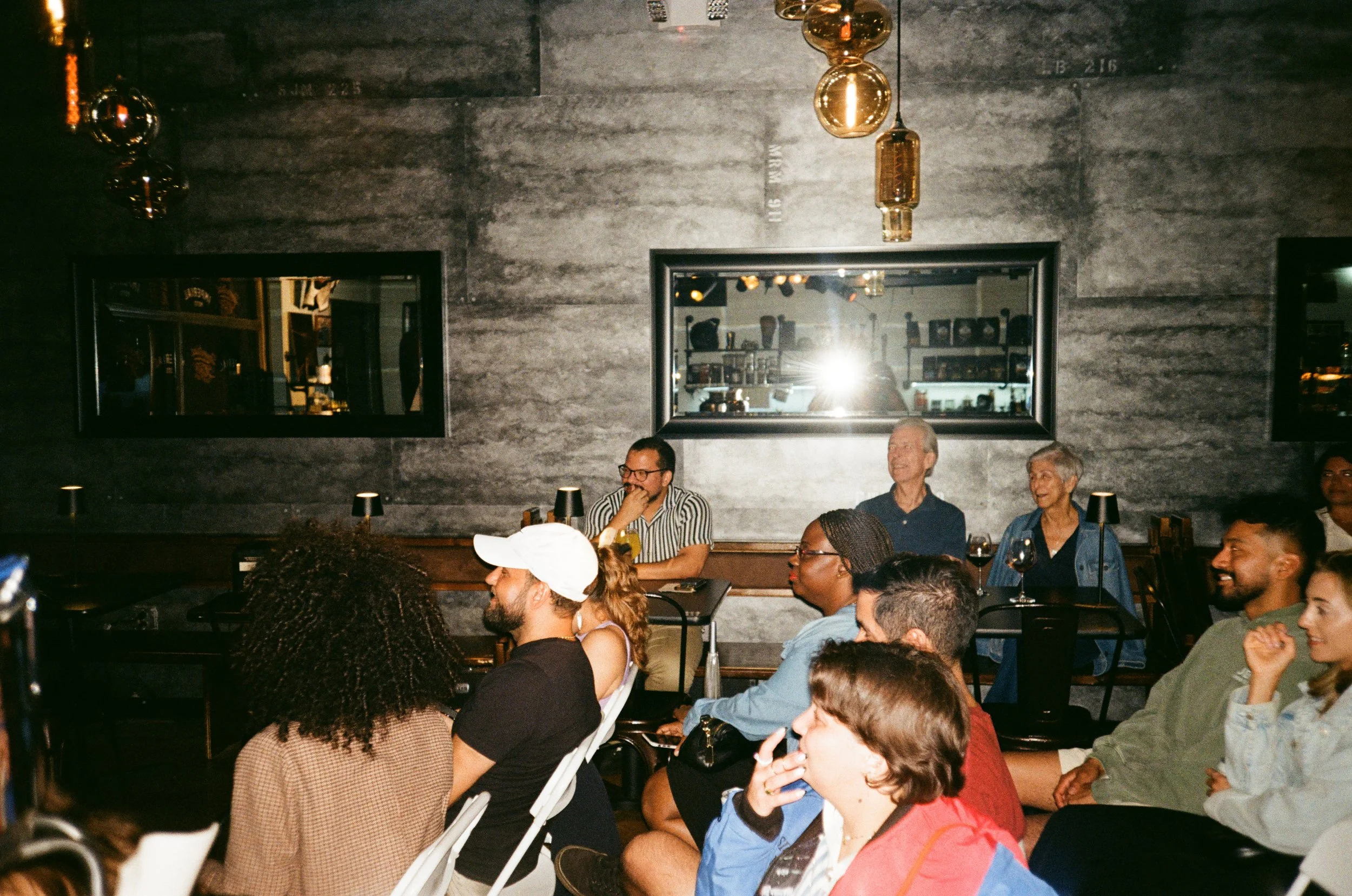 People sitting in a dimly lit room, listening and smiling, with wall-mounted mirrors and modern pendant lighting.
