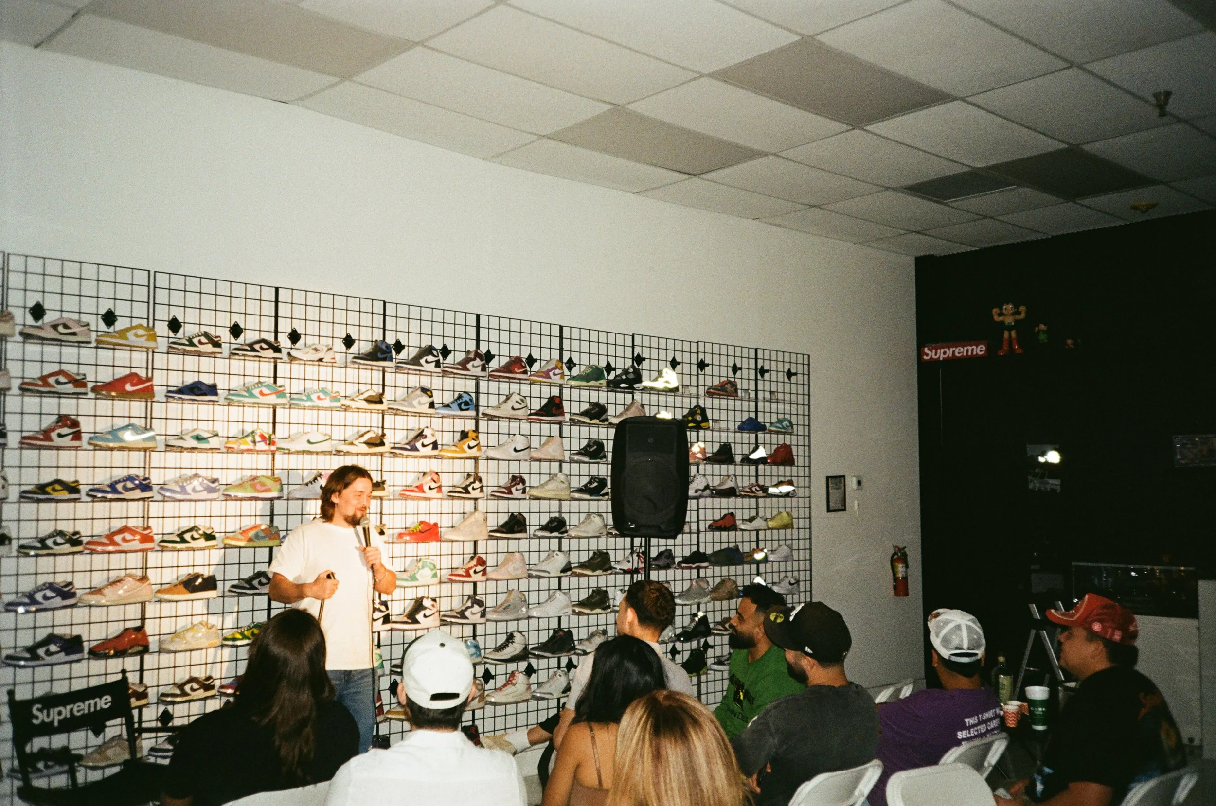 A man with long hair and a white shirt speaking into a microphone in front of a wall display of sneakers at a sneaker event. Several people seated and listening, some wearing caps, facing him.