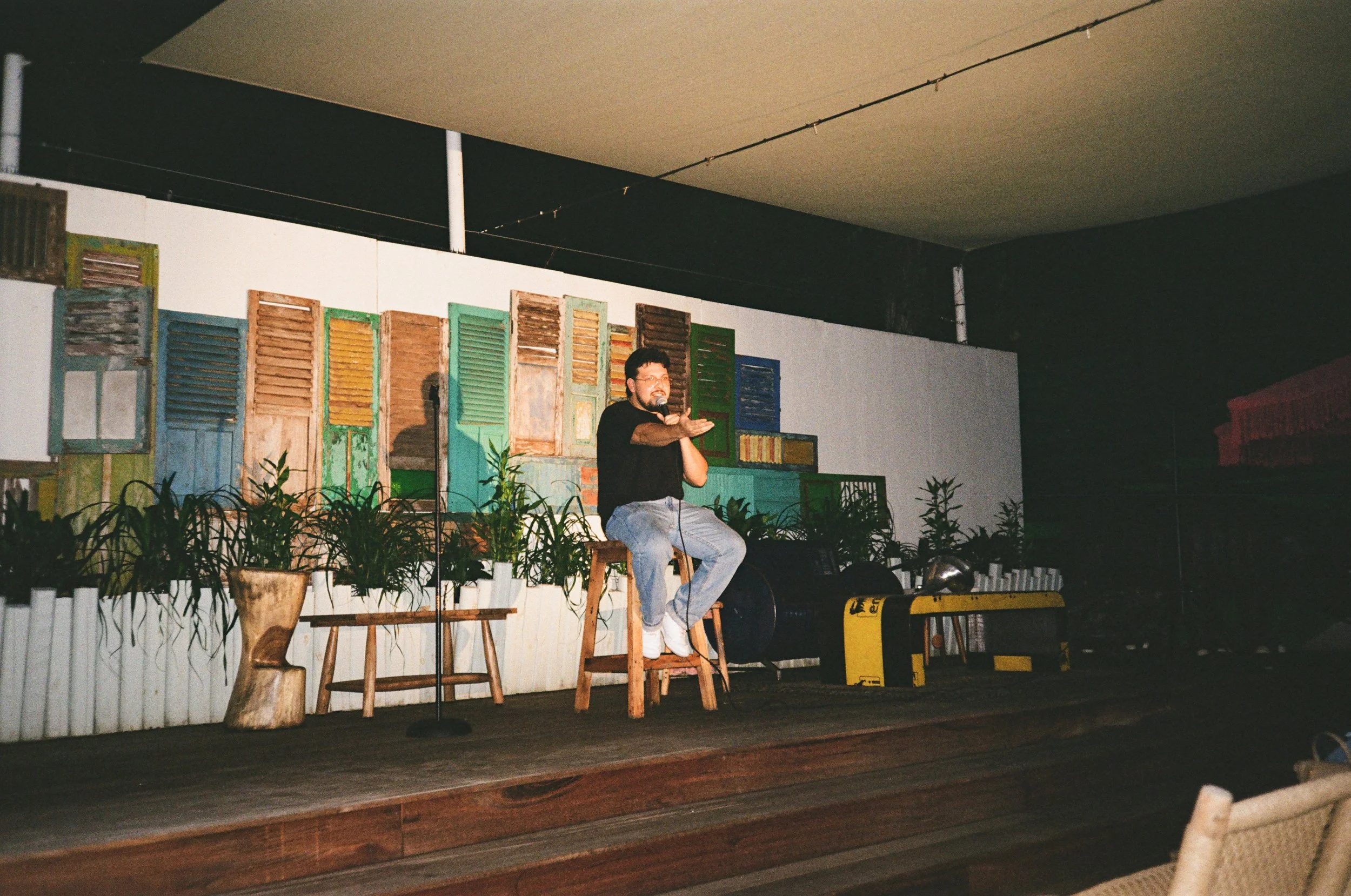 A man sitting on a stool on a stage, gesturing with his hands, performing in front of a backdrop of colorful, weathered window shutters and potted plants.