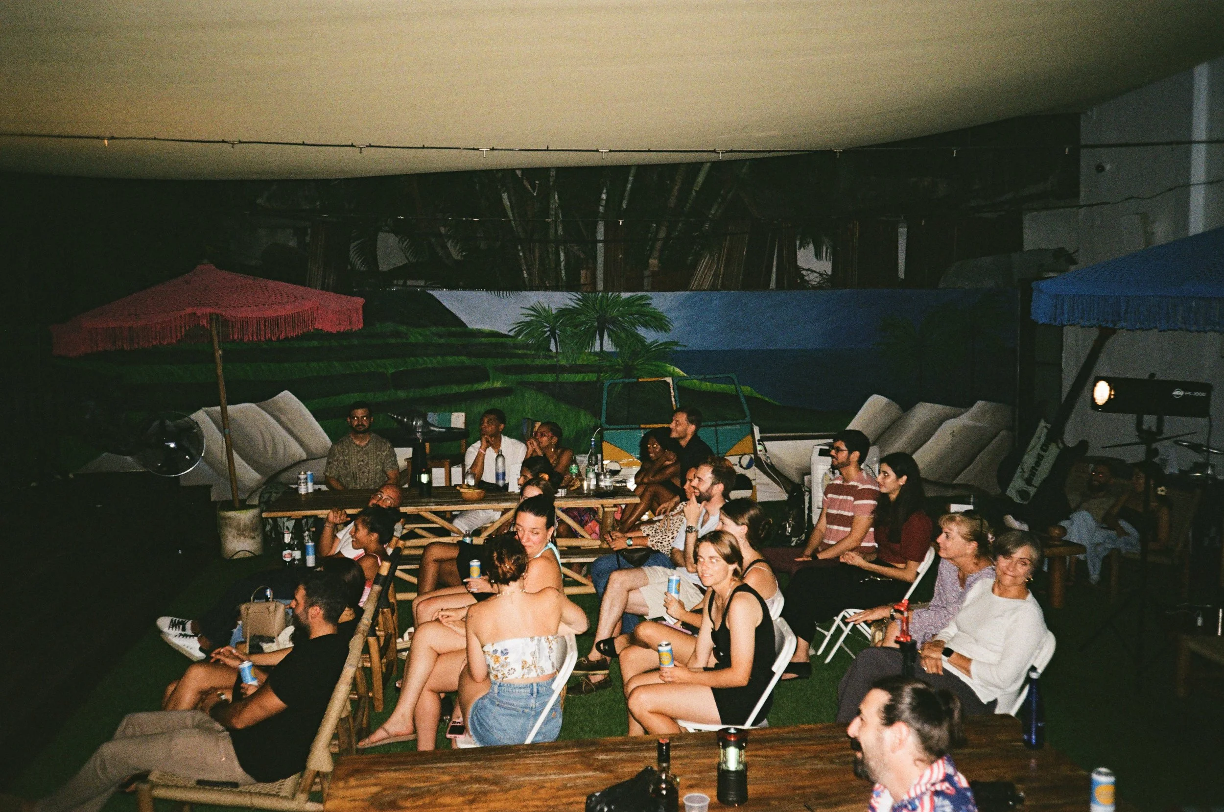 Group of people sitting on chairs and benches in an indoor space, watching a performance or presentation, with a mural of a landscape in the background.