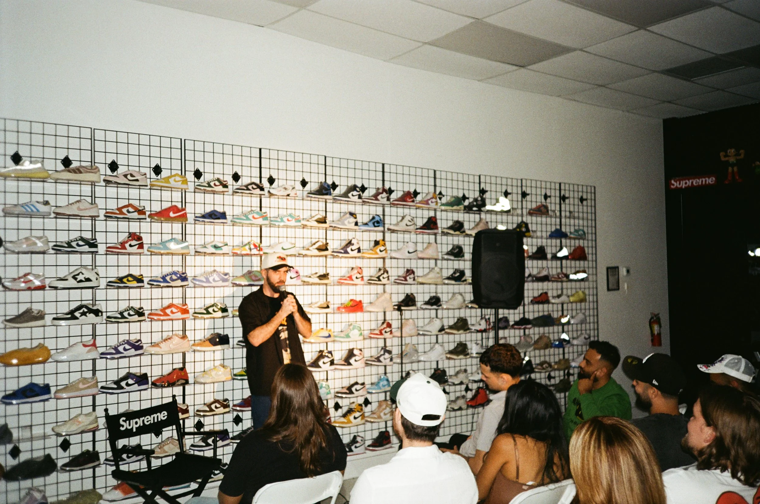 Man speaking into microphone in sneaker store with wall display of colorful sneakers behind him, audience seated and listening.