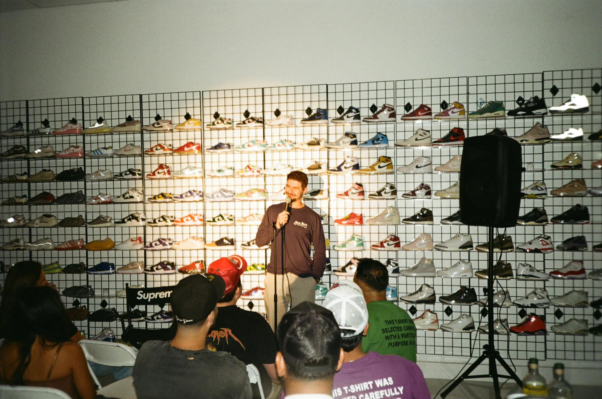 A man standing on stage in front of a wall filled with shoes, speaking into a microphone in front of an audience.