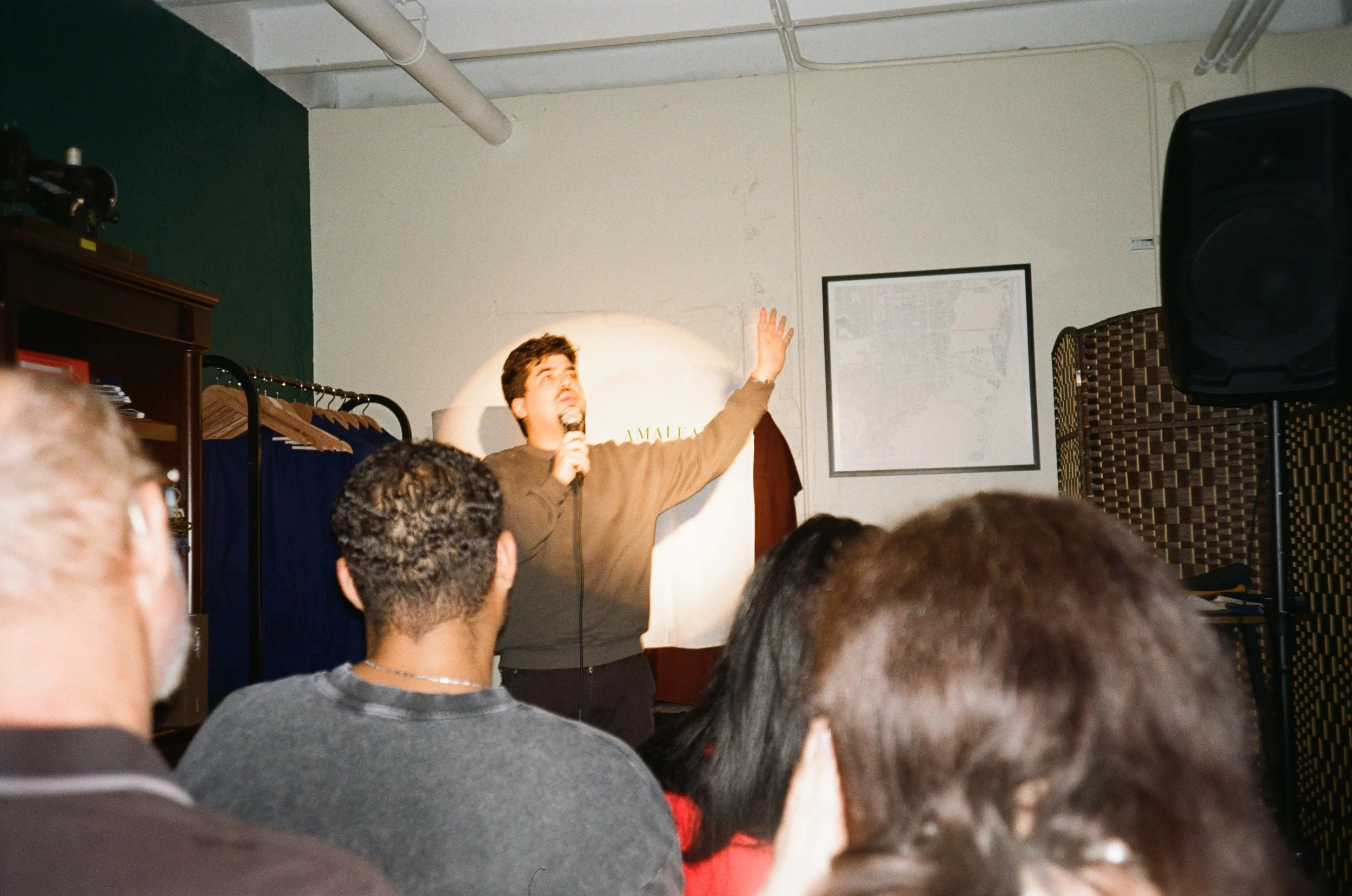 A man speaking into a microphone on a stage with a white screen behind him, gesturing with one arm raised, in front of an audience.