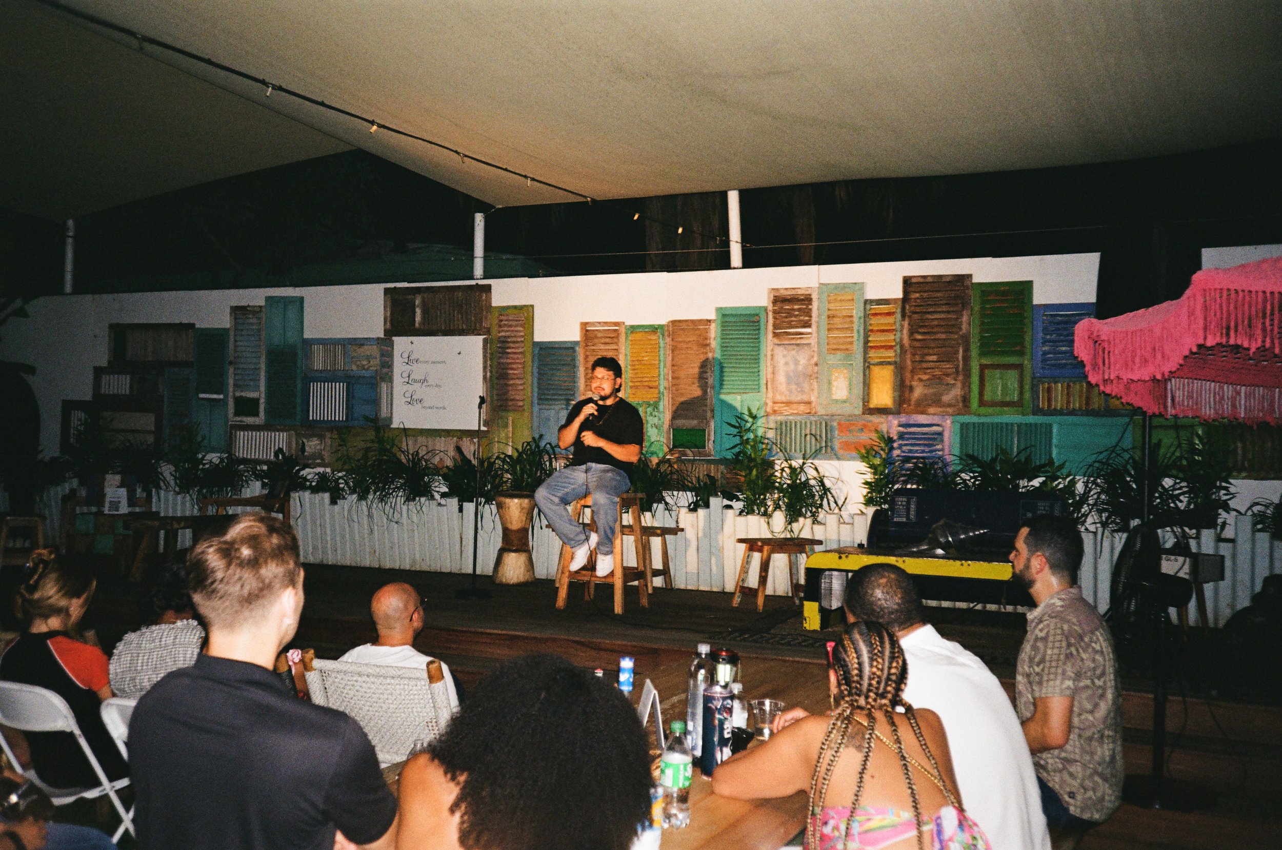 A man seated on a high stool with a microphone, speaking to an audience at an indoor event. The backdrop features a wall decorated with colorful, vintage window shutters and plants.