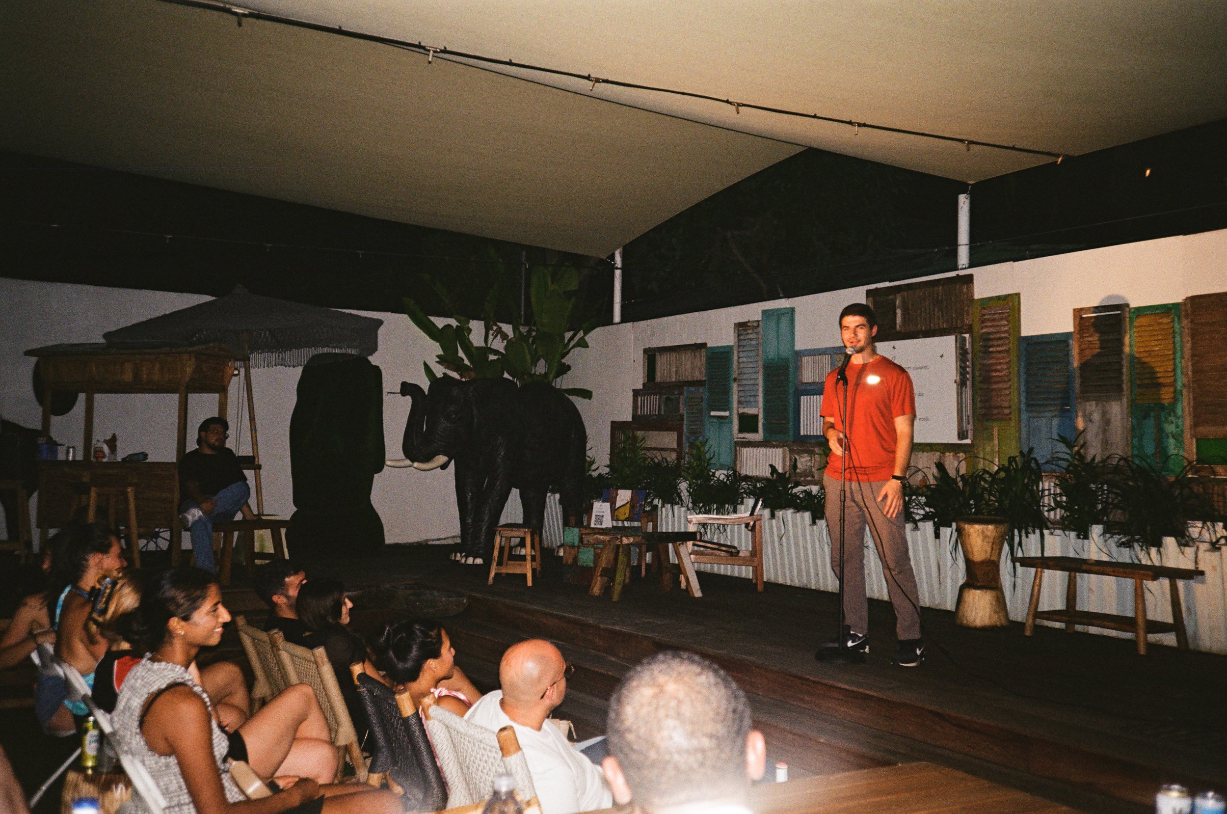 Man performing stand-up comedy on stage with audience watching in a tropical-themed venue decorated with colorful shutters, plants, and an elephant sculpture.