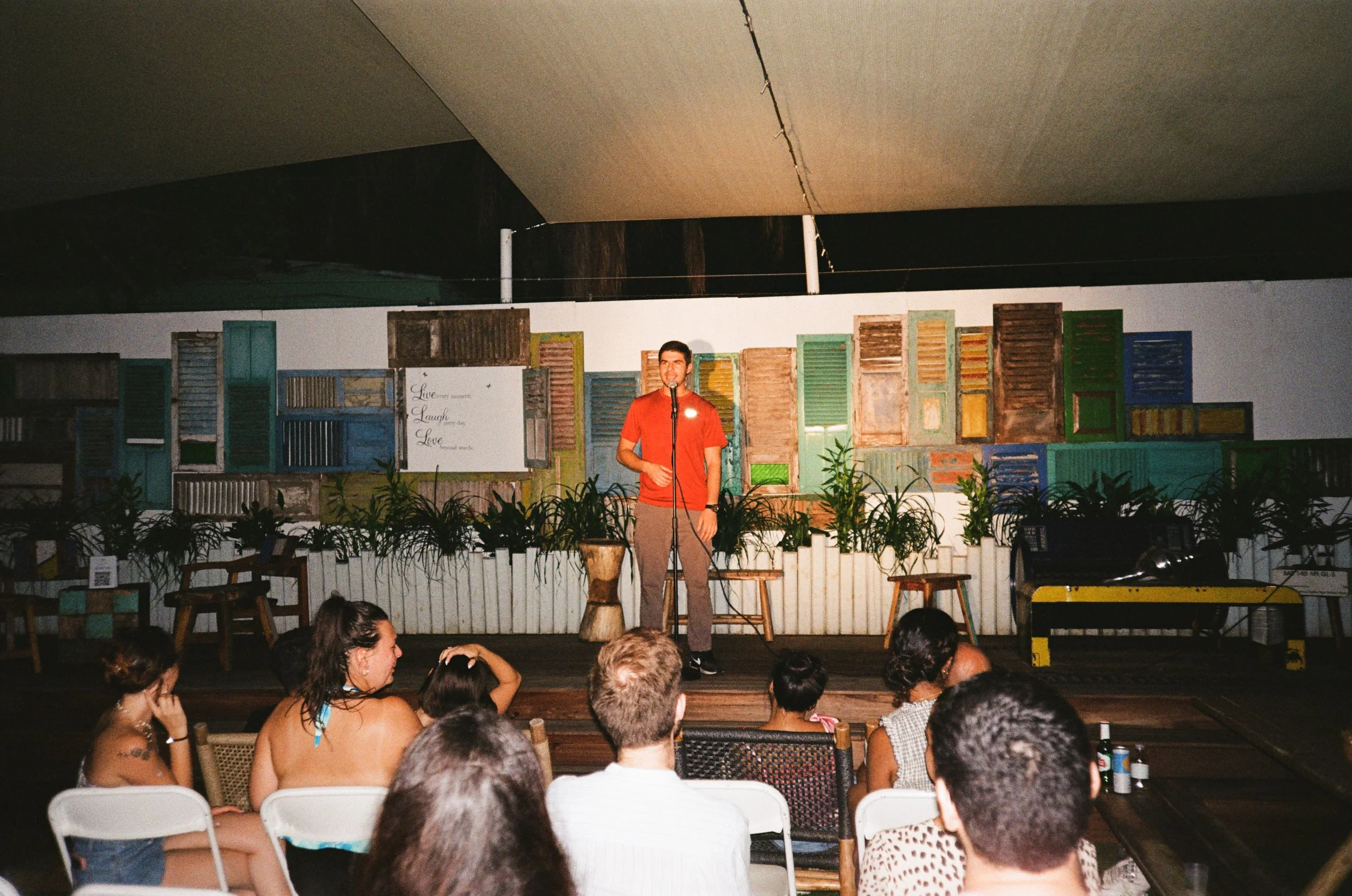 A man standing on a small stage, wearing a red shirt and speaking into a microphone, with an audience seated in front. The backdrop features a wall decorated with colorful vintage window shutters and a whiteboard with motivational words.