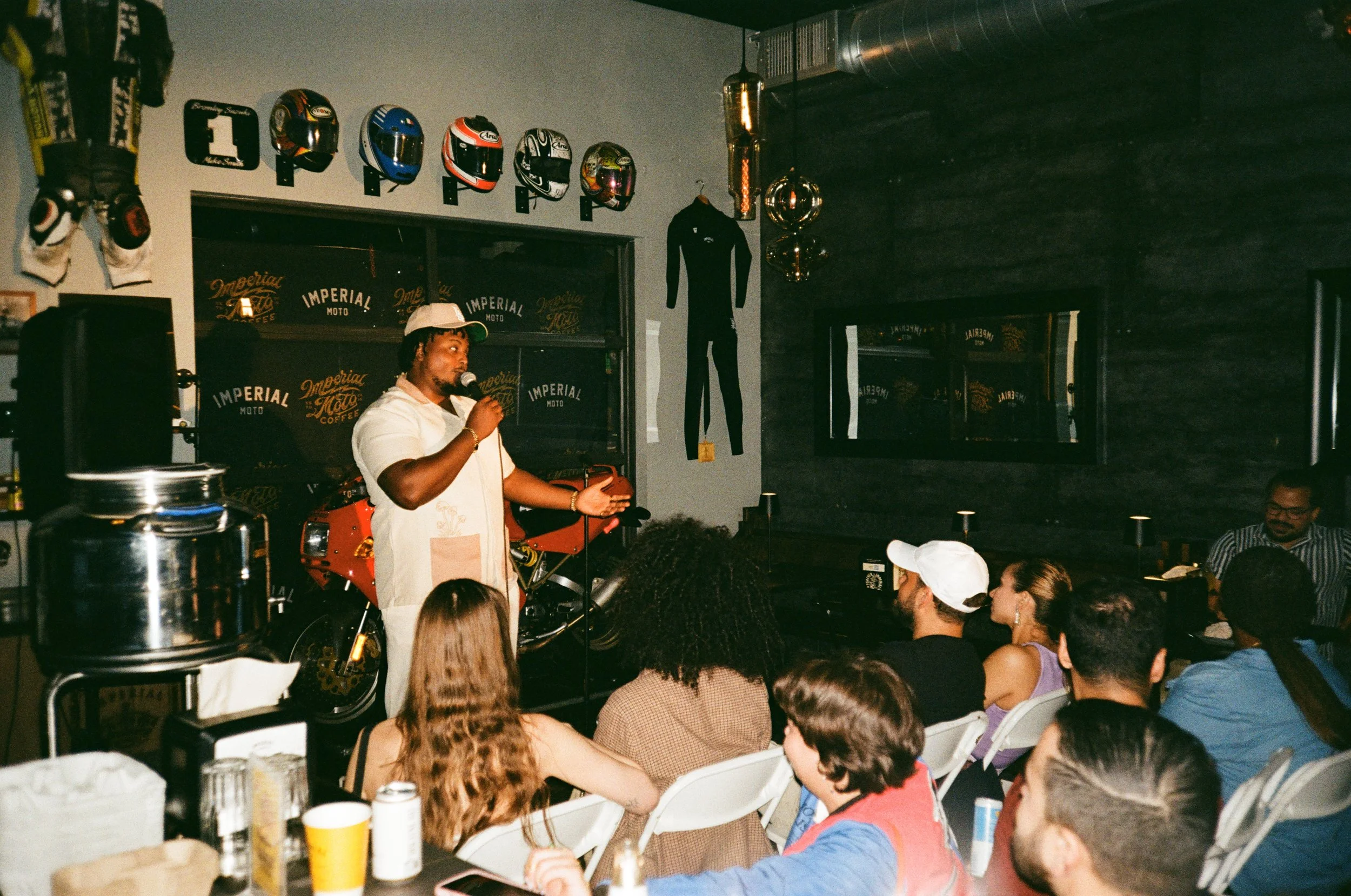 A man in a white shirt and white cap is speaking into a microphone to an audience seated in front of him in a dimly lit bar or cafe decorated with motorcycle helmets and racing memorabilia. There are several people of diverse backgrounds listening at