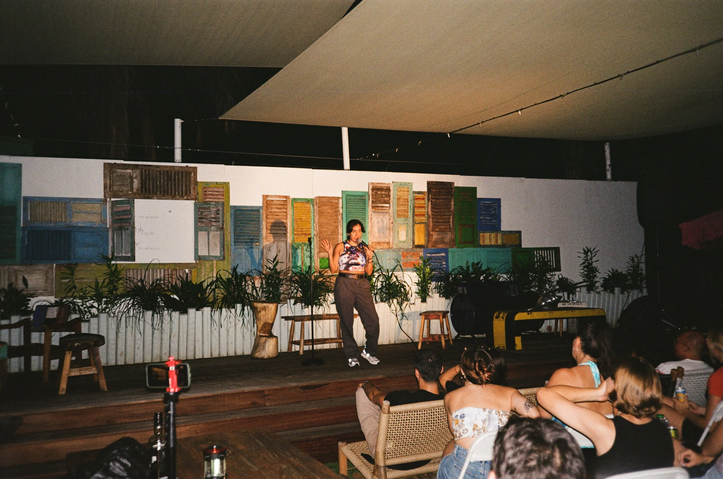 A woman performing on a stage in front of an audience, with a backdrop of colorful wooden shutters and plants, under a large ceiling canopy.