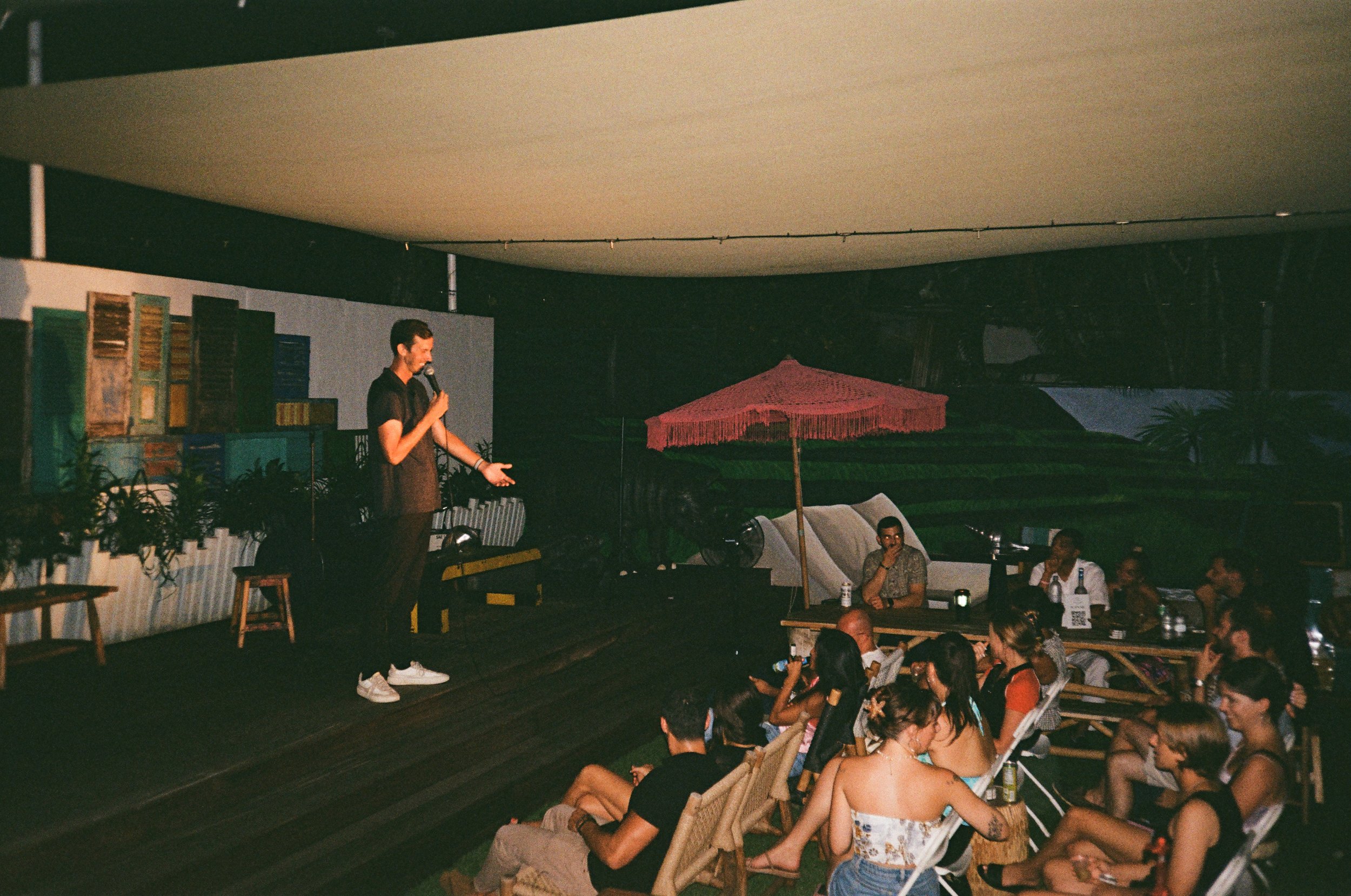 A man stands on a stage with a microphone, performing in front of an audience seated on chairs and benches. The stage has a backdrop of colorful bookshelves and plants, with a pink parasol and outdoor furniture nearby.