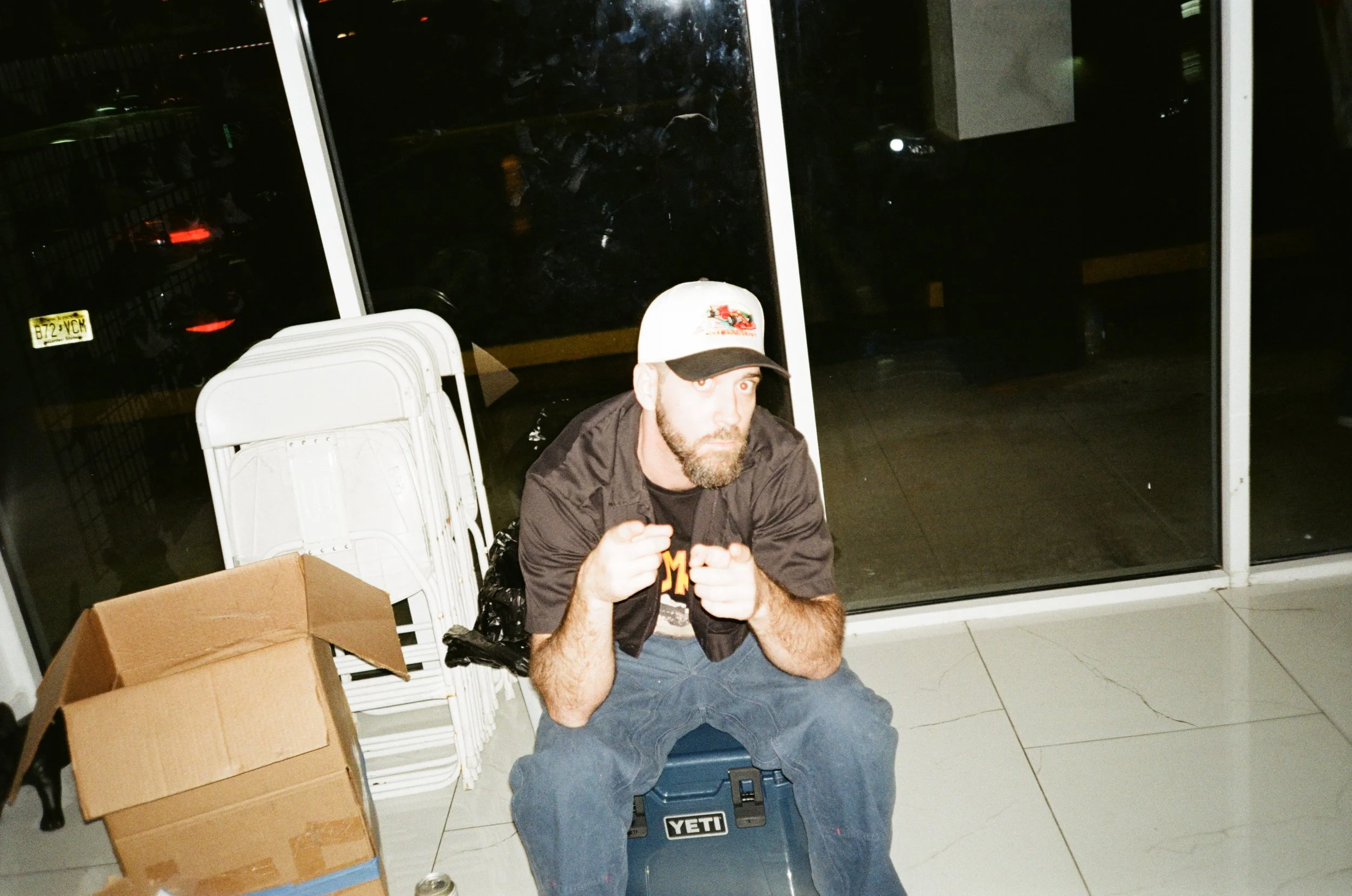 A man with a beard and a baseball cap sitting on a coolers, pointing at the camera with both hands, in an indoor setting with a large glass window behind him, stacked white folding chairs, a cardboard box, and a soda can on the floor.