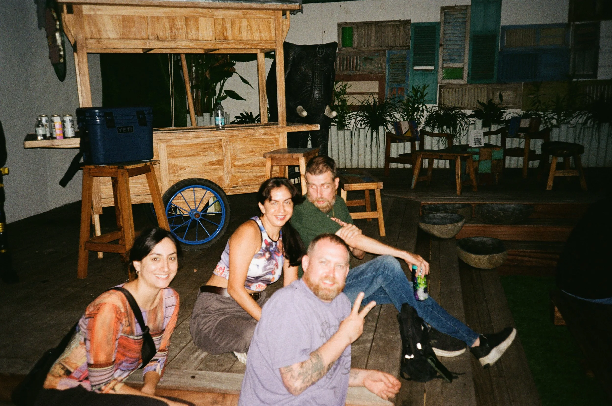 Four people sitting on a wooden floor in a casual indoor setting, smiling and posing for a photo. There are wooden chairs, tables, plants, and decorative elements in the background.