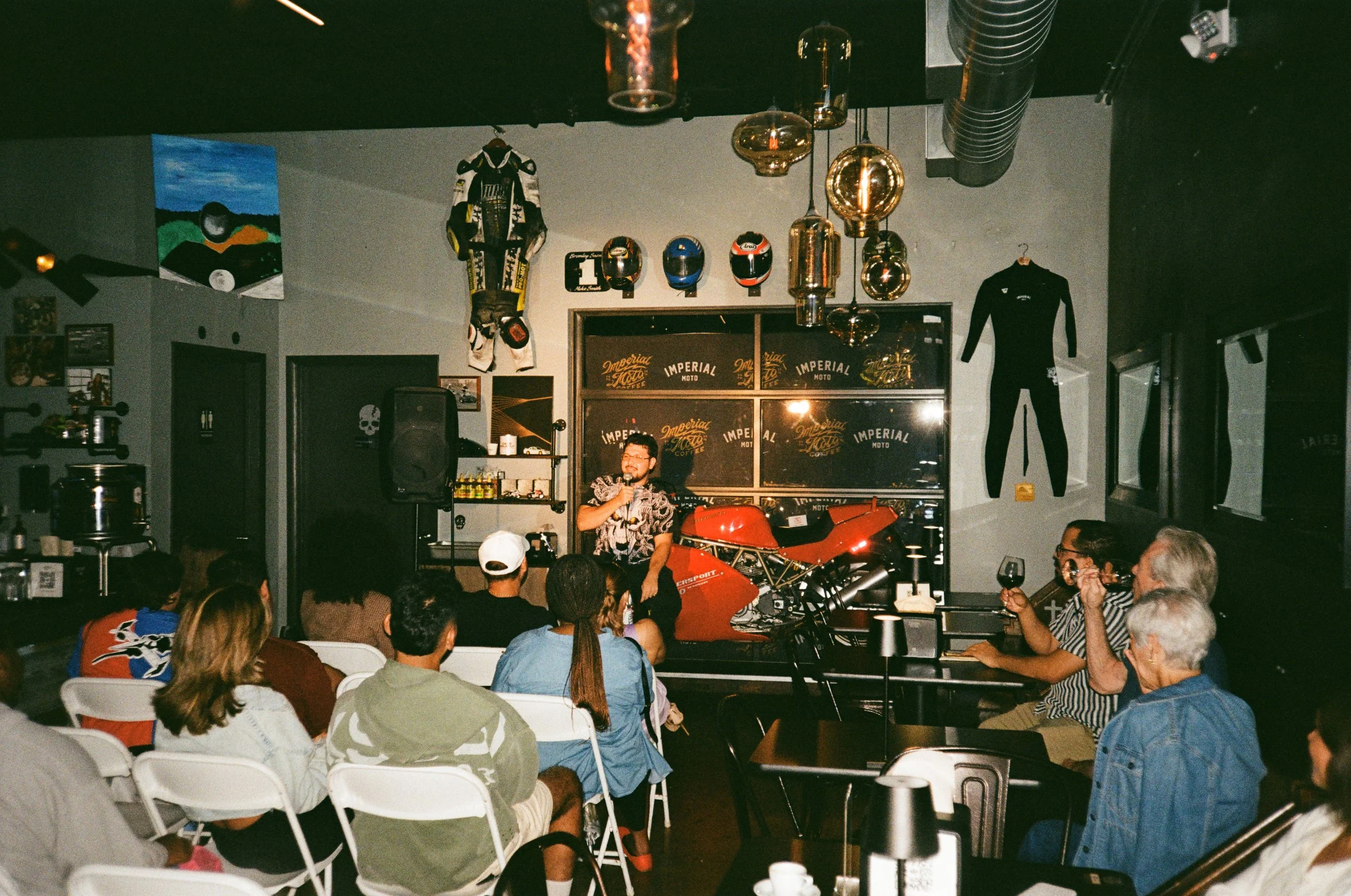 A man standing on a stage in a bar or club, speaking into a microphone to an audience seated on white chairs, with motorcycle helmets and racing suits as decorations.