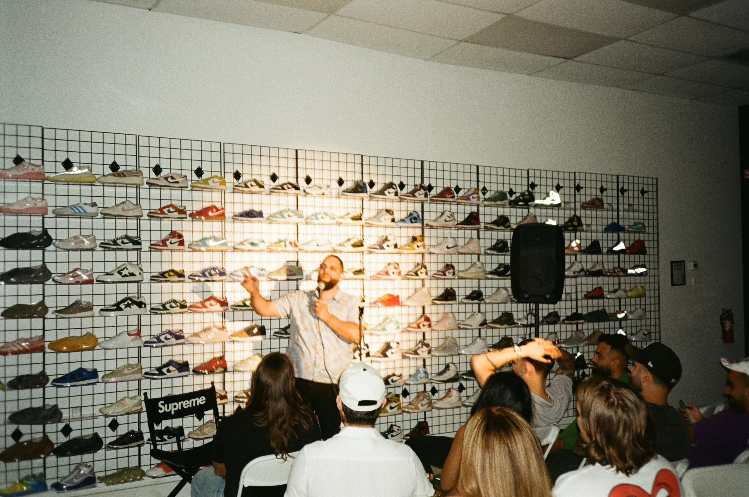 A man with a microphone speaking to an audience seated in front of a wall decorated with numerous colorful sneakers on a grid display