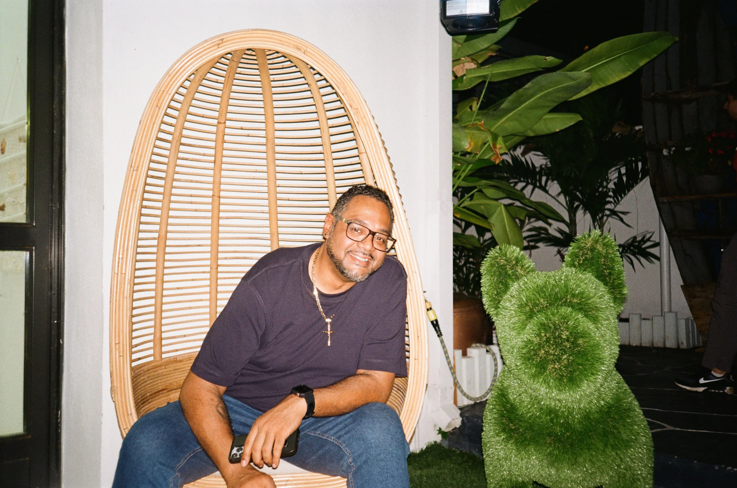 A man sitting on a rattan egg-shaped chair, smiling, holding a smartphone, wearing glasses, a black T-shirt with a cross necklace, and a wristwatch, outdoors at night with large green plants and a planter shaped like a rabbit nearby.