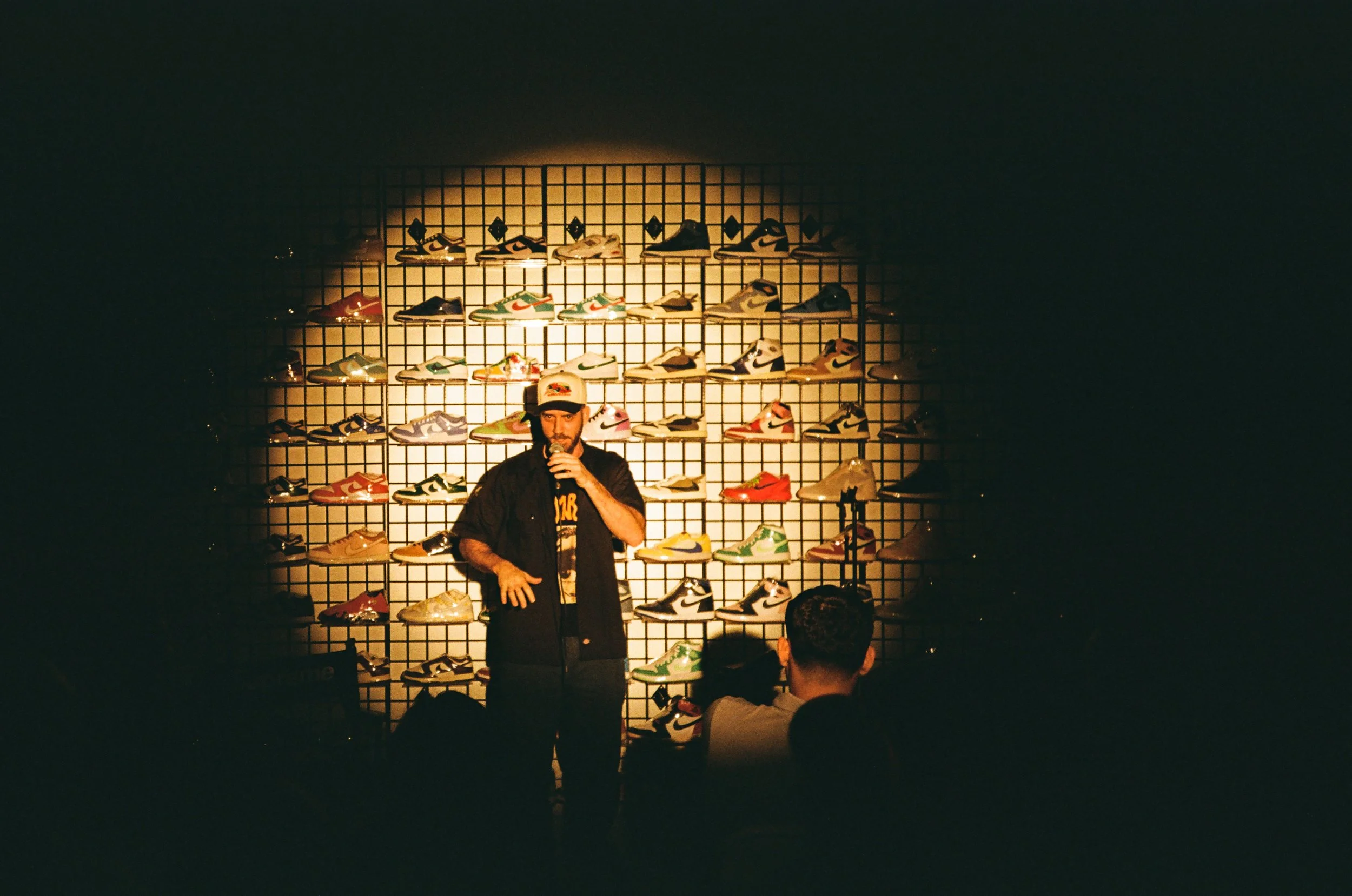 A man with a microphone standing in front of a display wall of various colorful sneakers, with audience members seated in front.