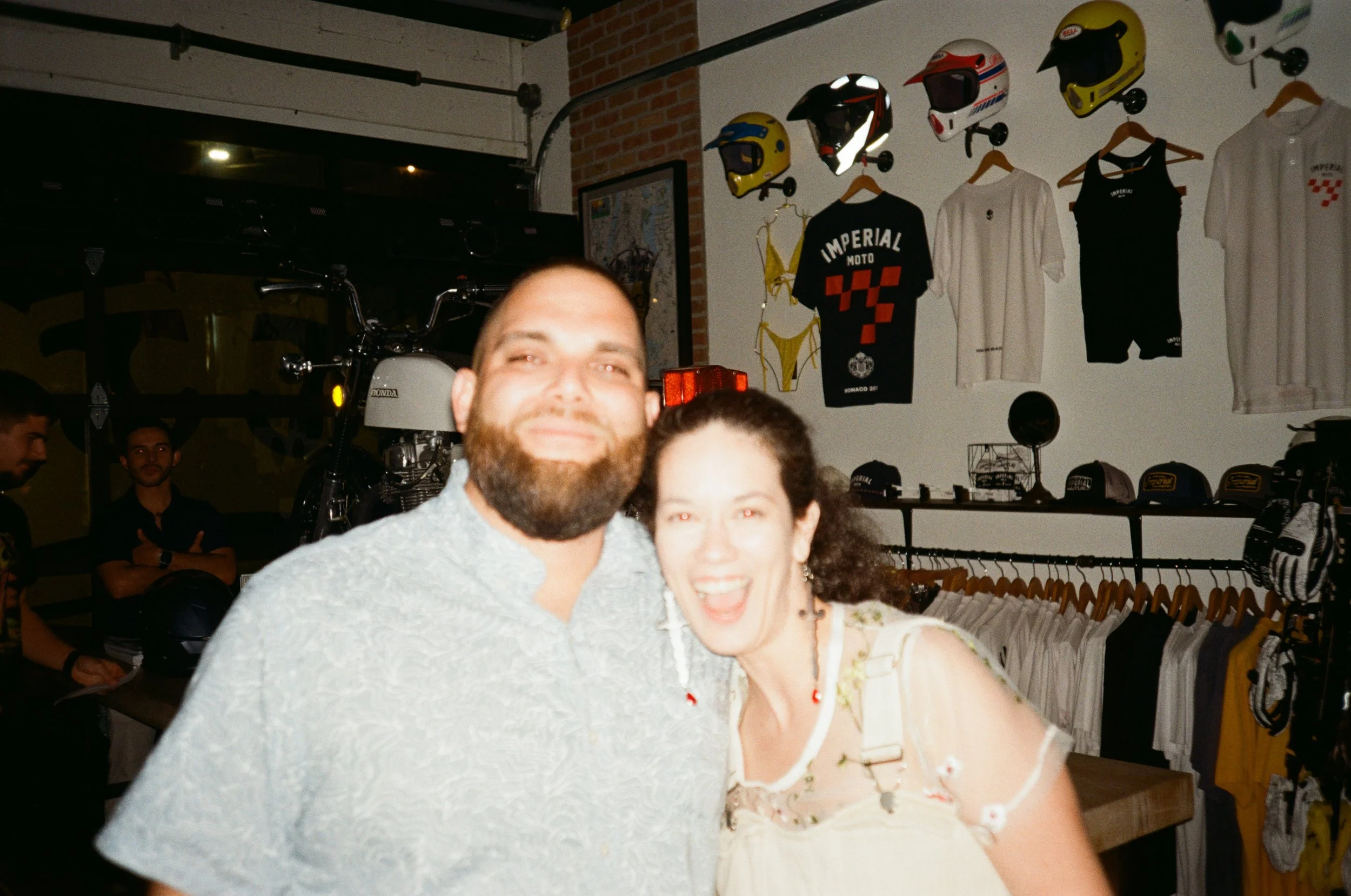 A smiling man with a beard and a woman with curly hair and smiling, posing inside a motorcycle shop or store with helmets and apparel on the wall behind them.