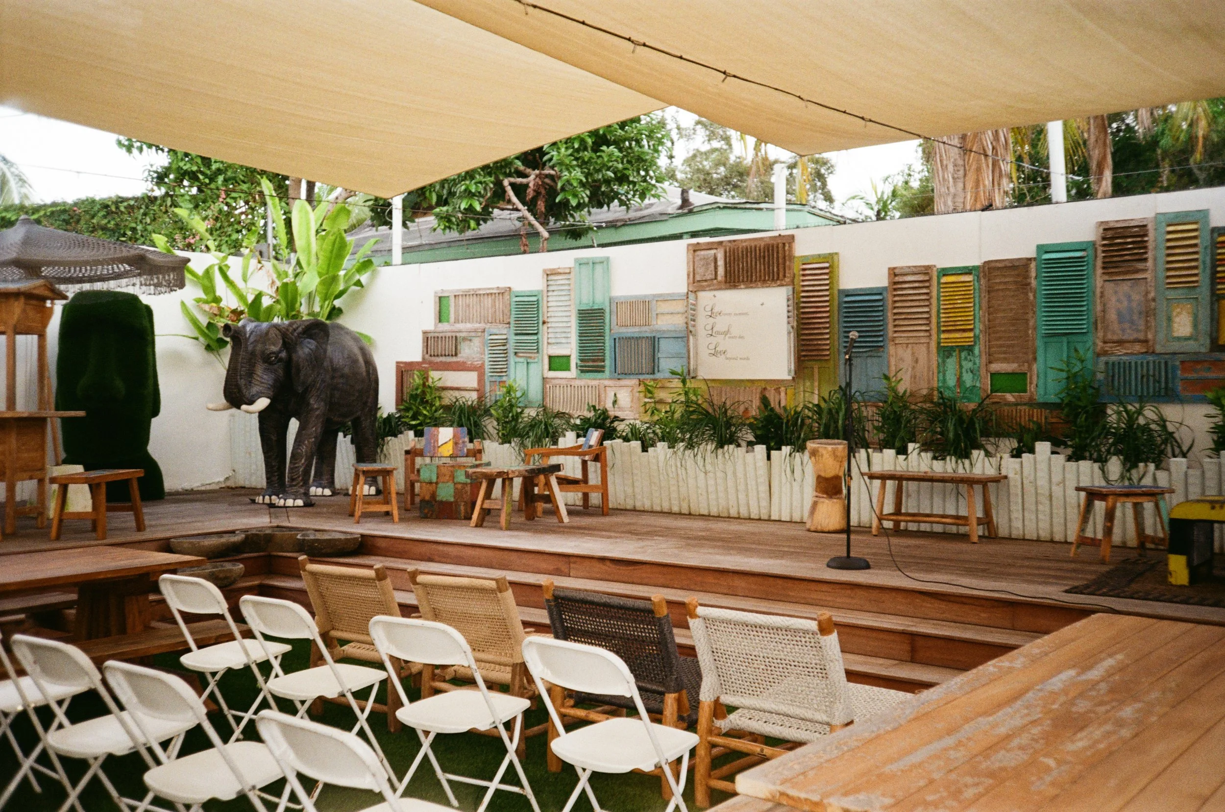Outdoor stage with a large elephant sculpture and a colorful display of reclaimed window shutters, surrounded by chairs and plants, under a beige shade cloth.