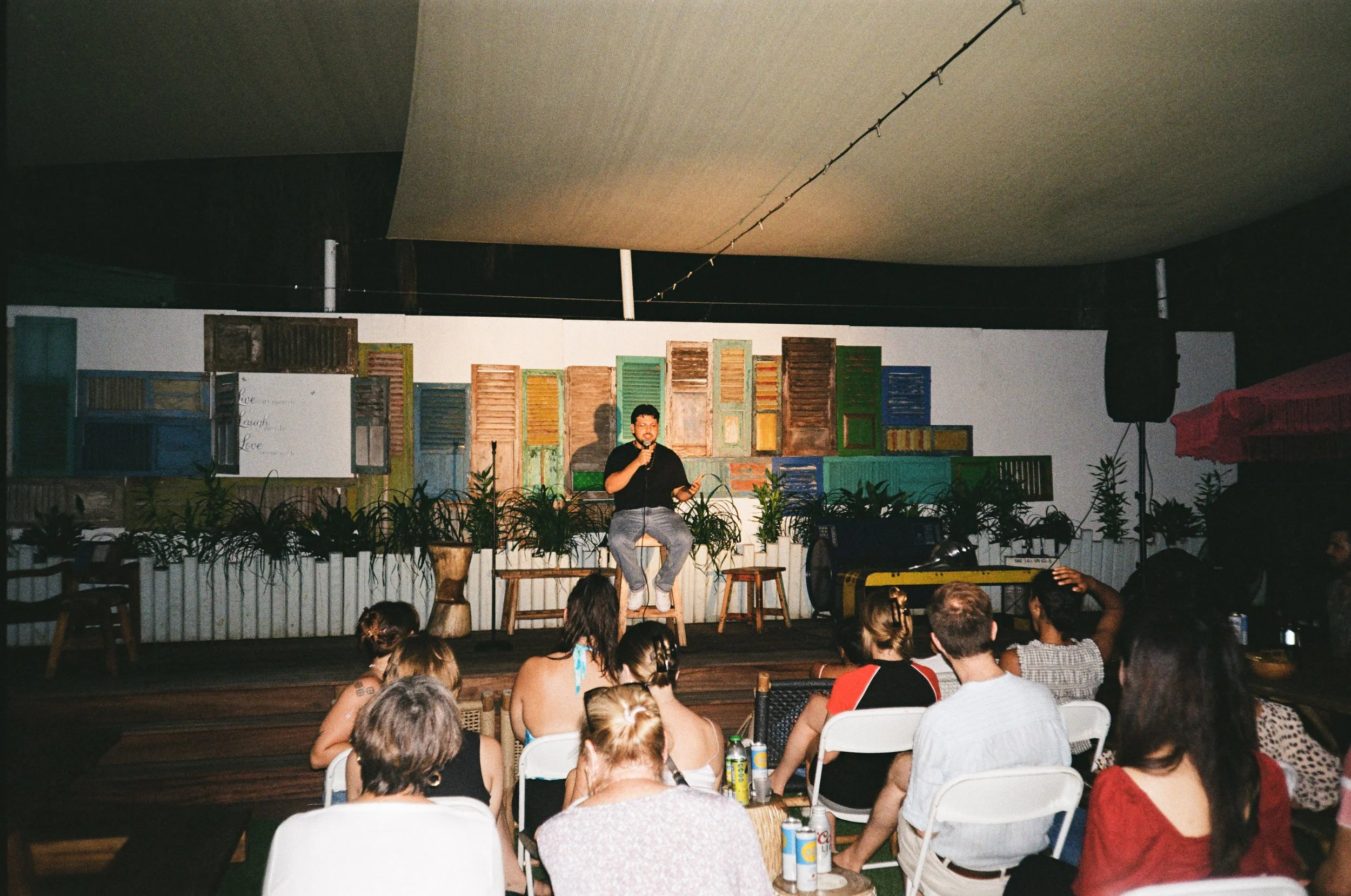 A man performing stand-up comedy on stage in front of an audience at night, with colorful wooden window shutter decorations on the wall behind him.