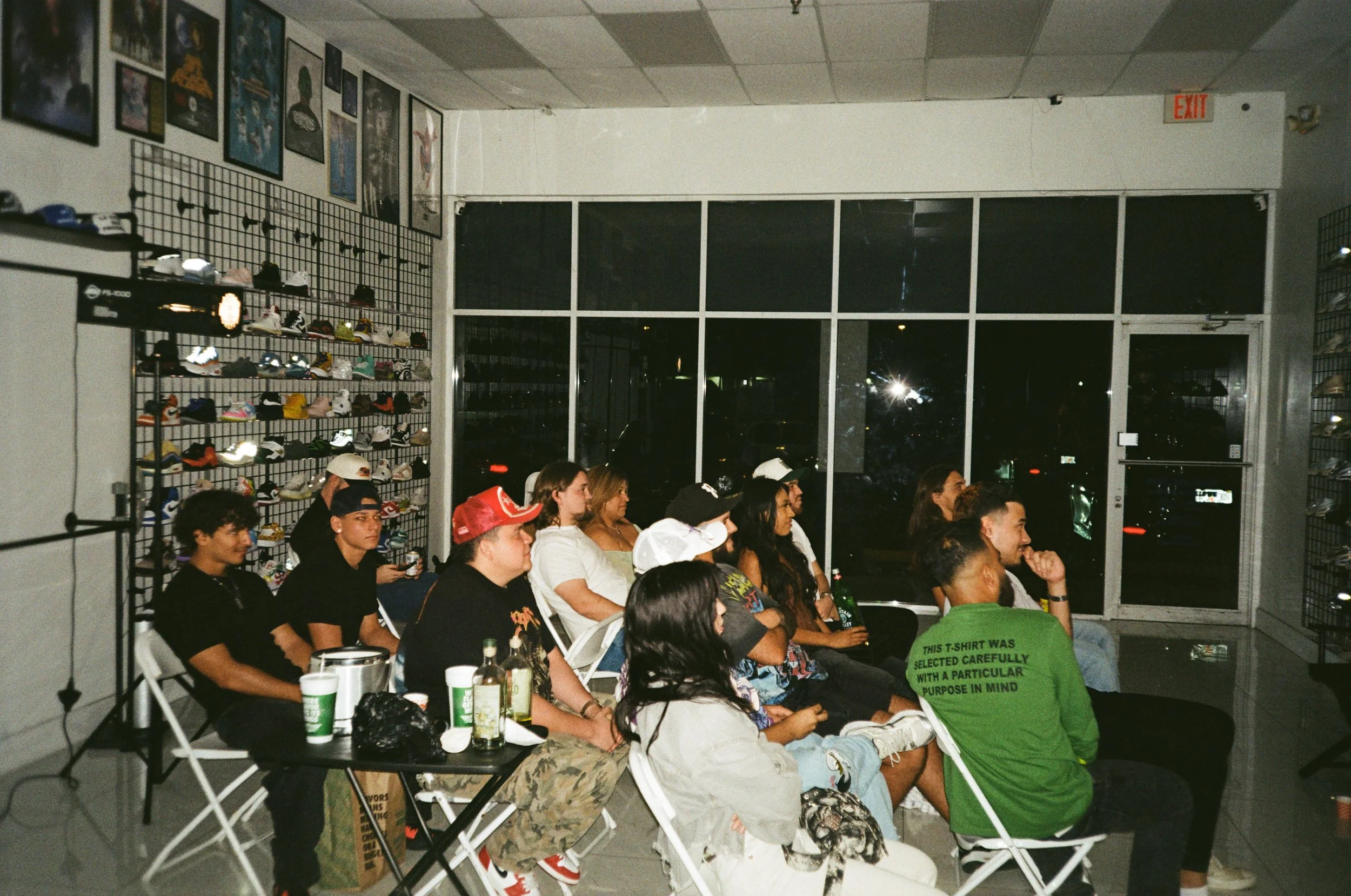 A group of people seated in chairs inside a retail store, watching a screen. The store has shoes displayed on wire grid walls and posters on the wall. It appears to be nighttime outside.