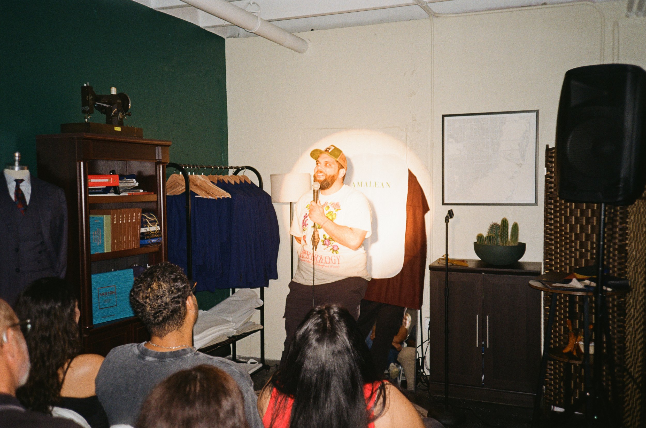 A man with a beard and a cap, standing on a stage with a microphone, speaking to an audience in a cozy room with green and white walls. The background includes a clothing rack with suits and jackets, a bookshelf, and decorative items like cacti.