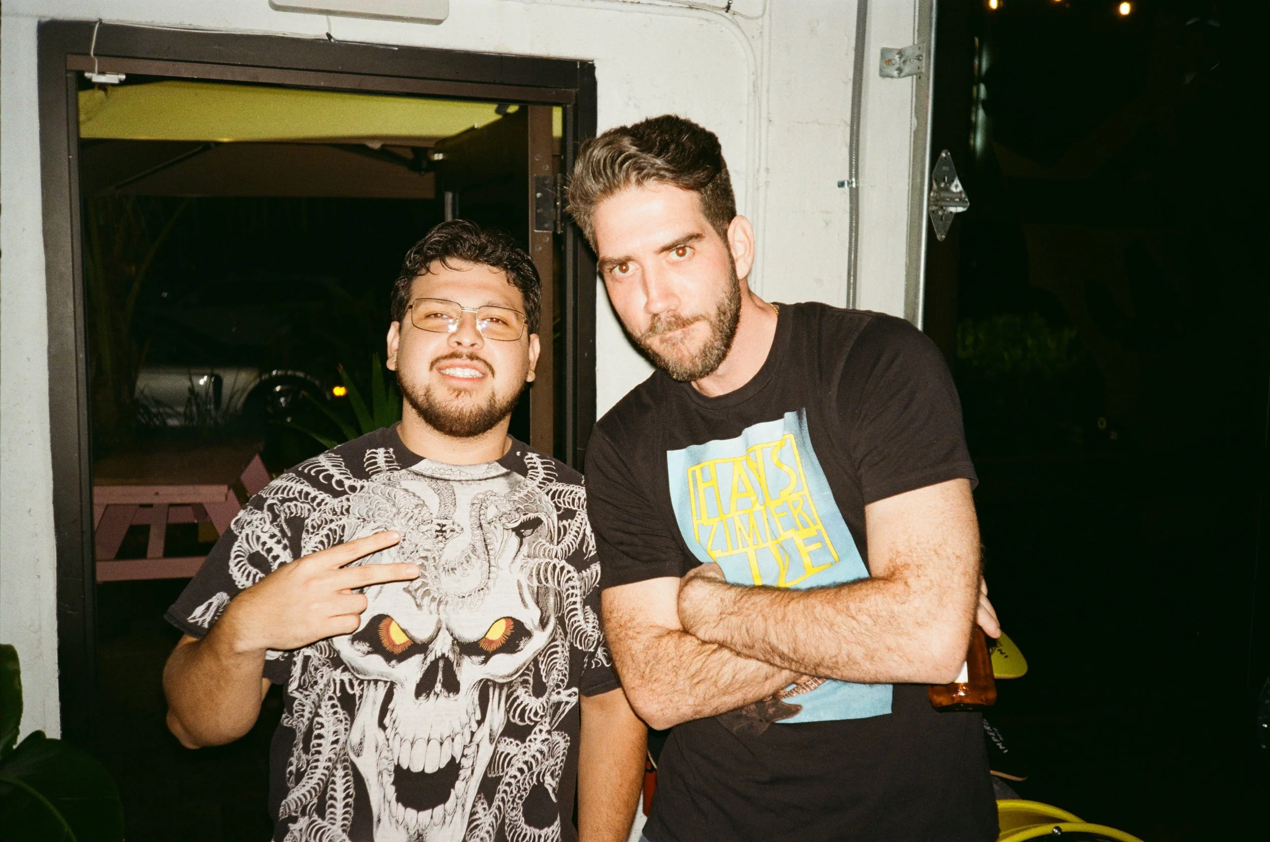 Two men standing together outside at night. The man on the left wears glasses and a shirt with a skull and lion design, making a peace sign. The man on the right has a beard and wears a black T-shirt with yellow and blue text, holding a bottle in his