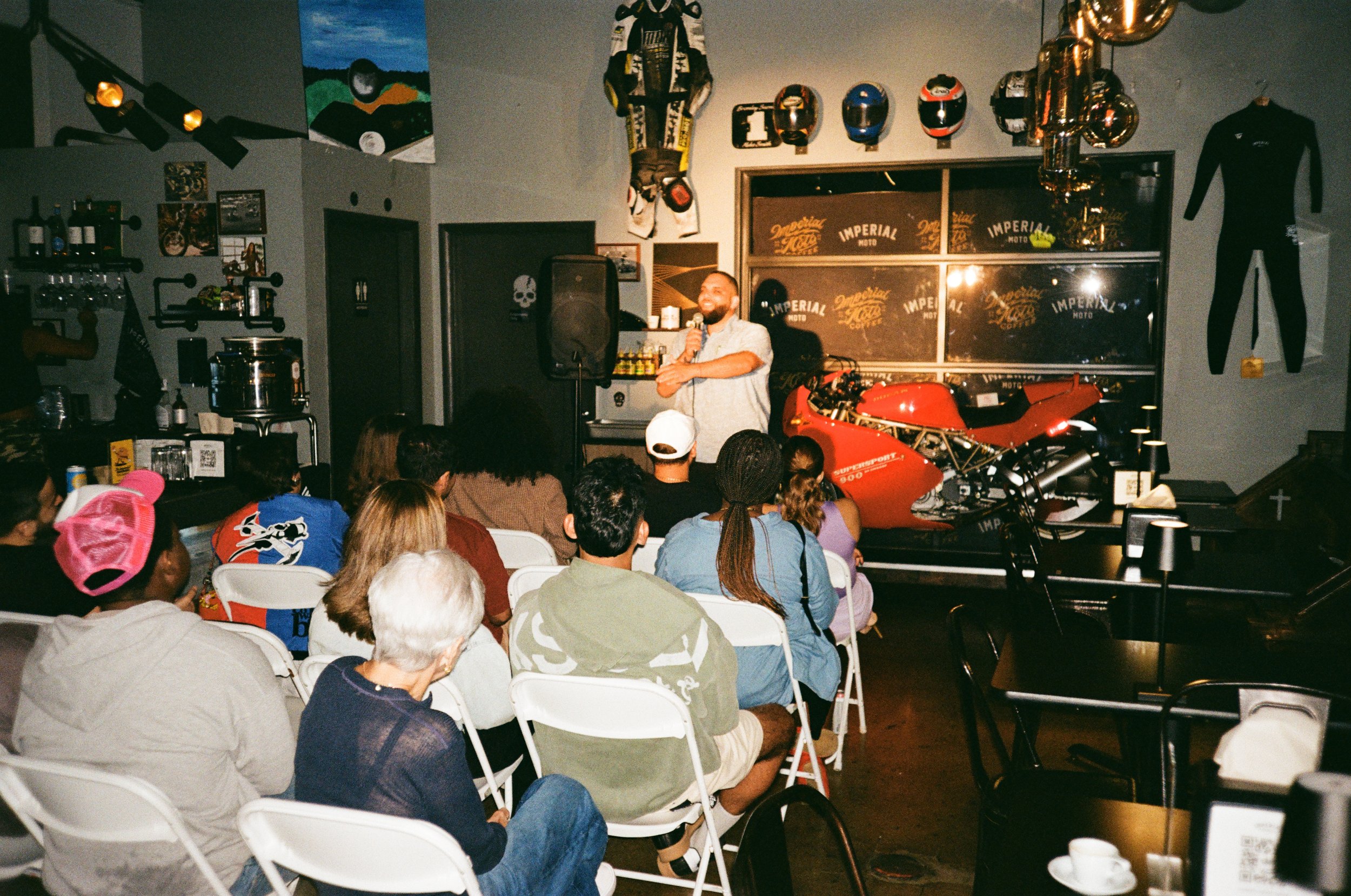 A man is giving a speech or presentation to an audience in a casual venue decorated with motorcycle memorabilia, helmets, and racing gear. The audience is seated on white chairs, and some people are wearing casual clothing and accessories.