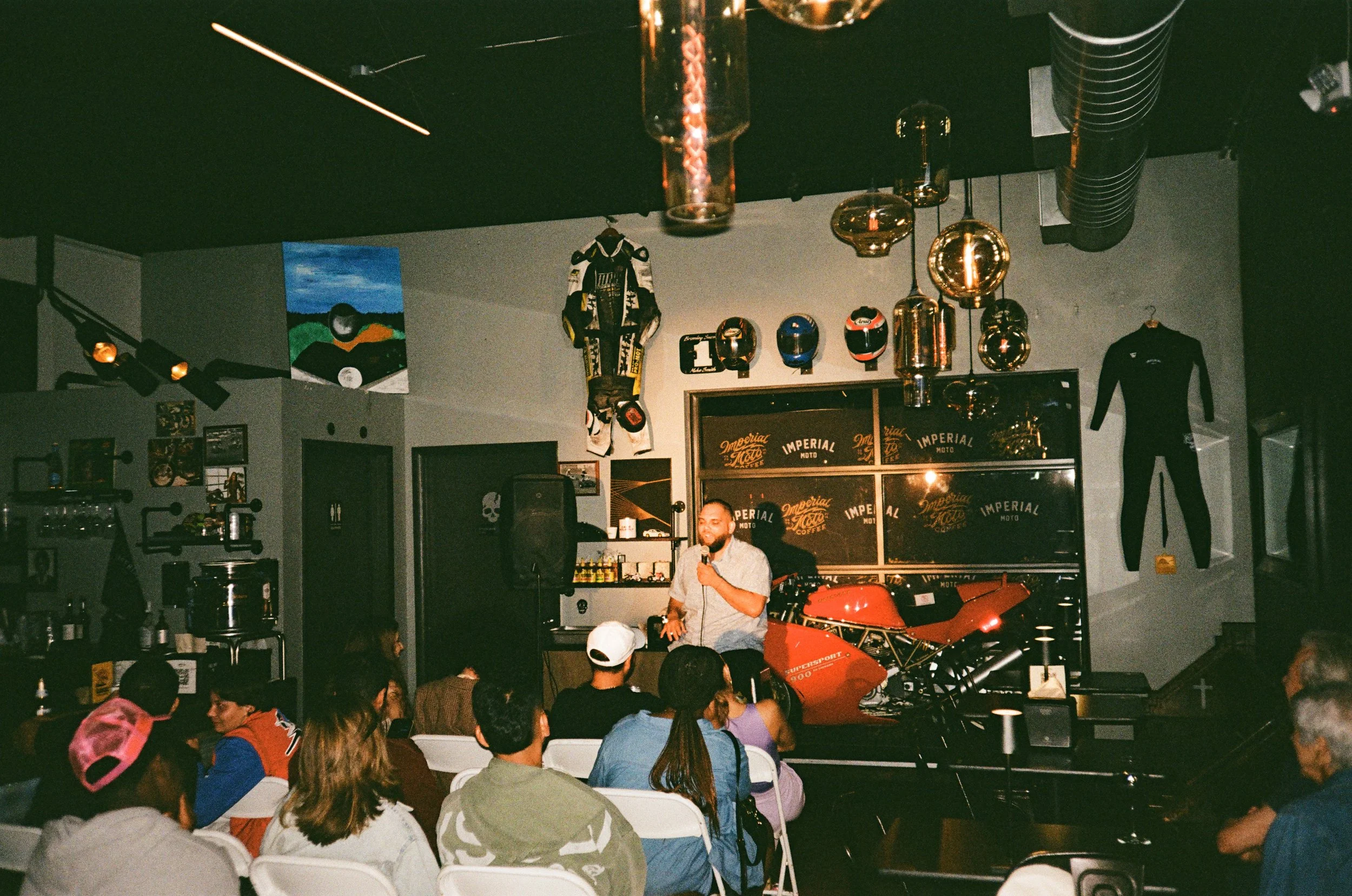 A man standing on a stage speaking into a microphone in front of an audience. The stage has a red motorcycle, motorcycle helmets, racing suits, and various memorabilia. The room has dim lighting with decorative hanging lights and artwork on the walls