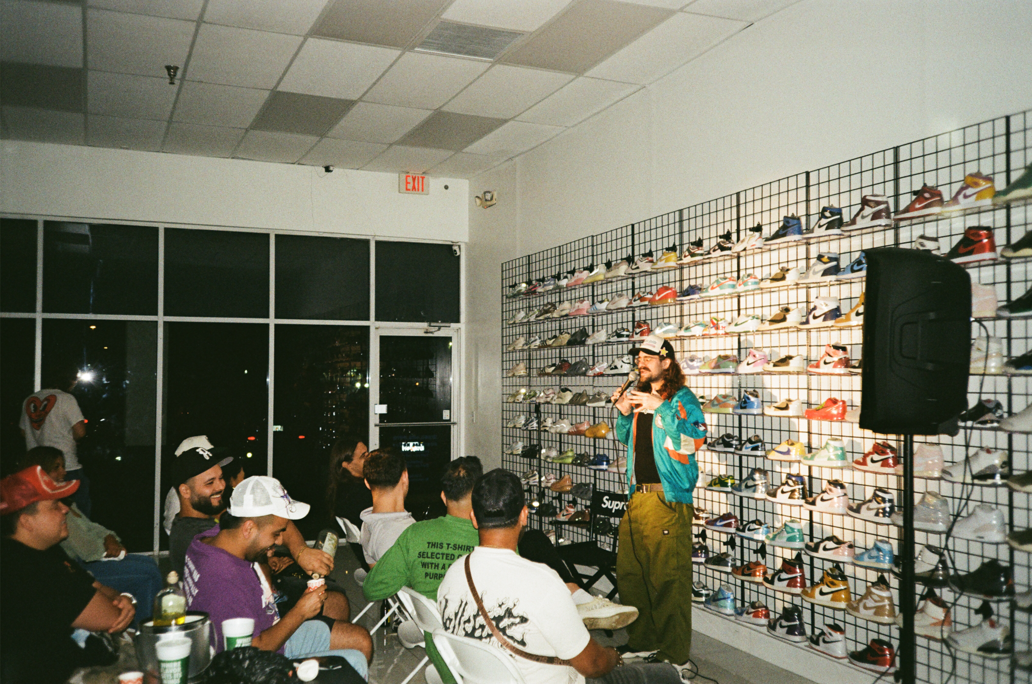 A person is speaking into a microphone in front of a wall of colorful sneakers displayed on a grid at a sneaker event, with an audience seated and watching.